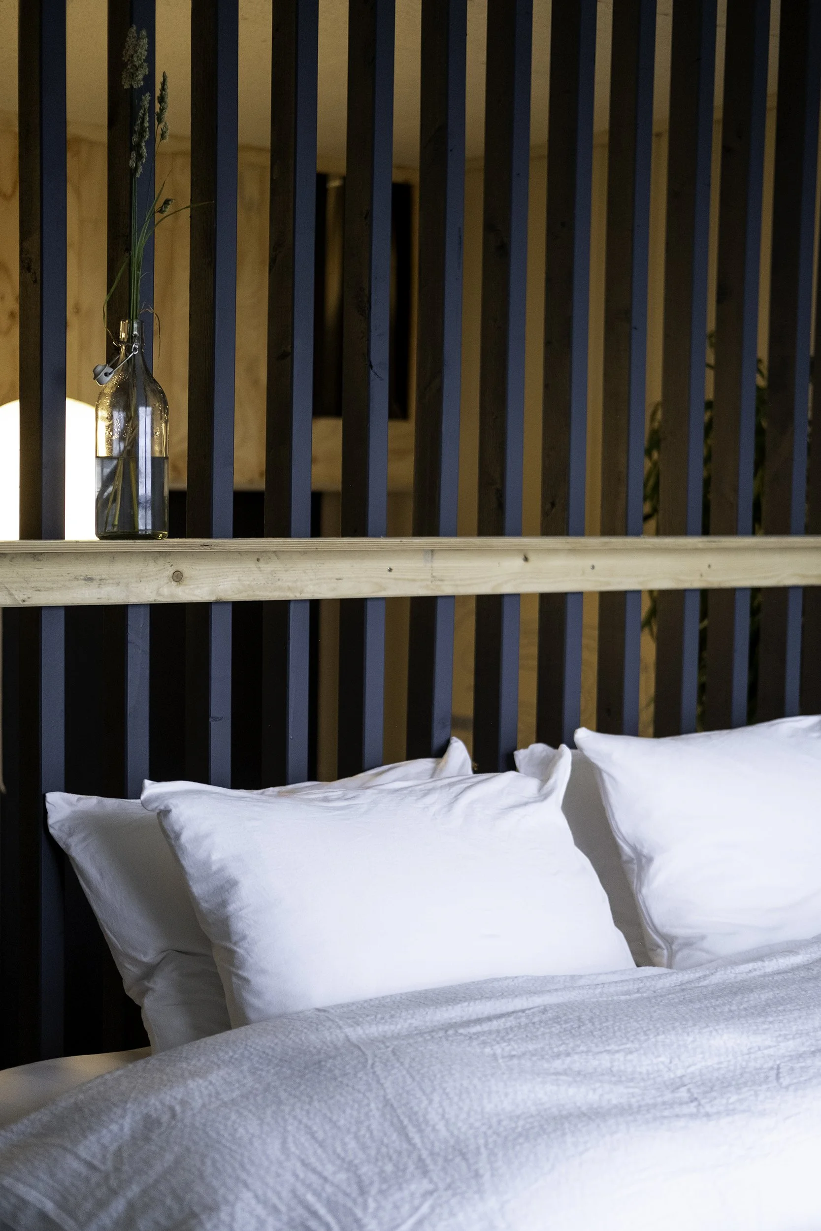 A bedroom with white pillows and bedding, a wooden headboard with vertical black slats, and a glass bottle with a single flower attached to a string on a shelf.