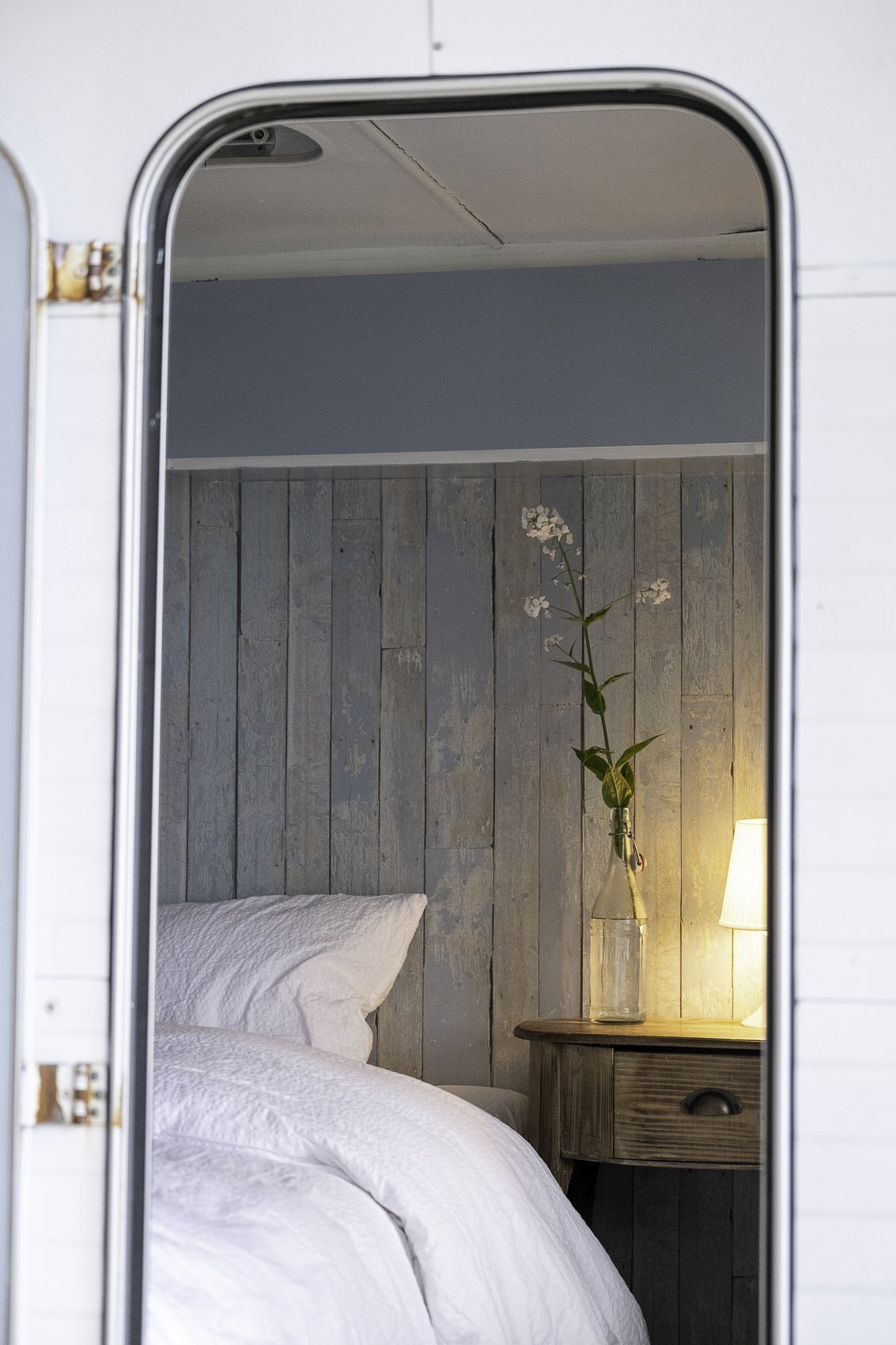 Inside a cozy bedroom with a white bed and pillows, a wooden nightstand with a glass vase and a tall plant, and a lamp illuminating the scene, seen through a doorway or mirror