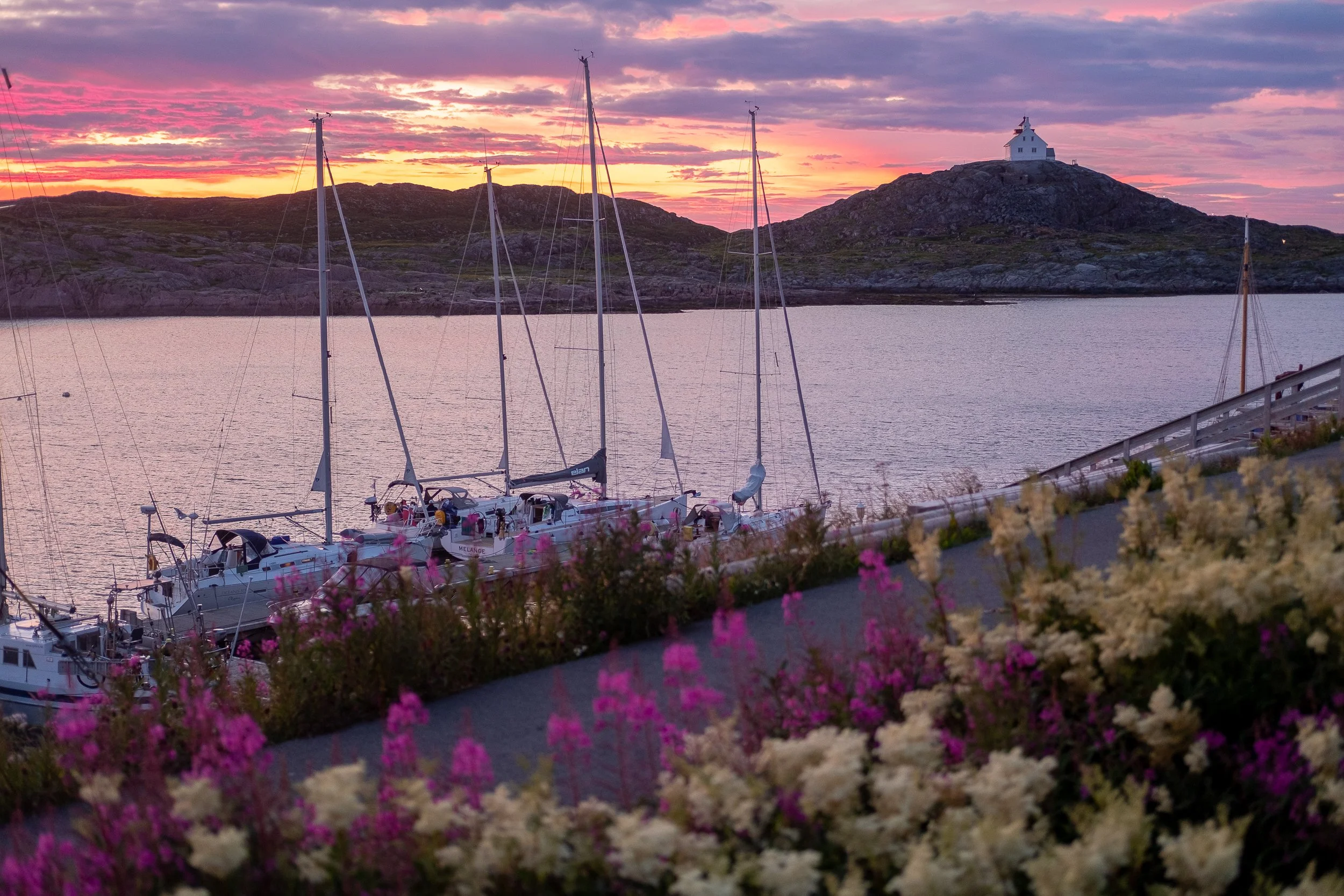 Sailboats docked along a harbor at sunset with pink and purple sky and a small white chapel on a hill in the background, surrounded by flowers and a railing in the foreground.