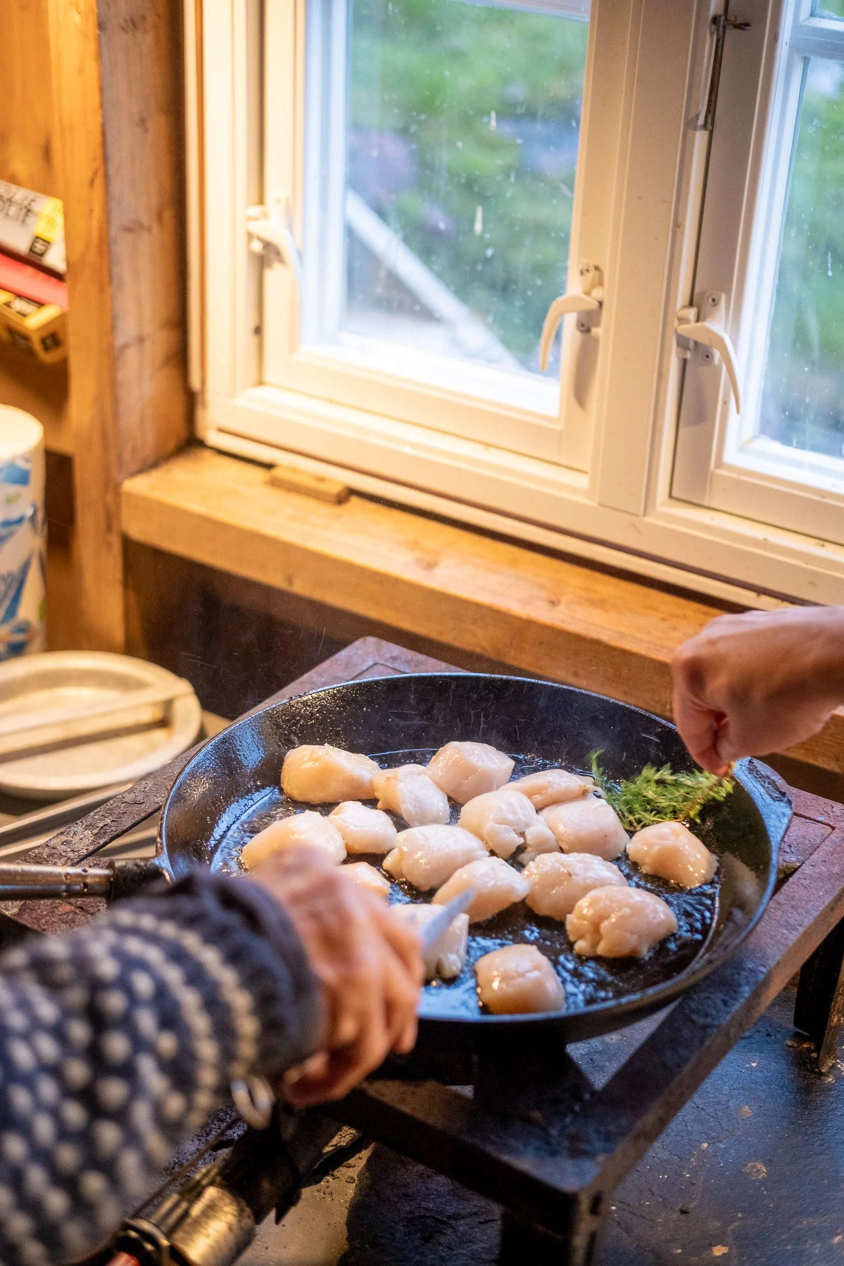 Person cooking scallops in a cast iron skillet on a stovetop in a rustic kitchen with a window showing an outdoor view.