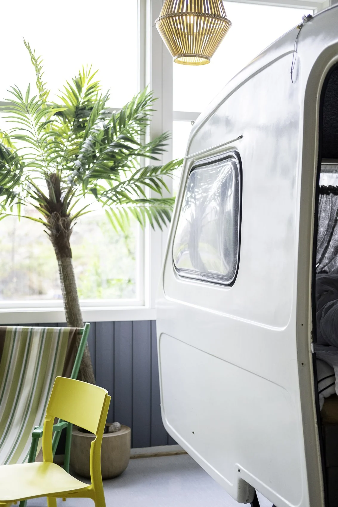 Interior of a room with a large window, a potted plant, a yellow chair, a lounge chair with striped fabric, and a partially visible white camper trailer.
