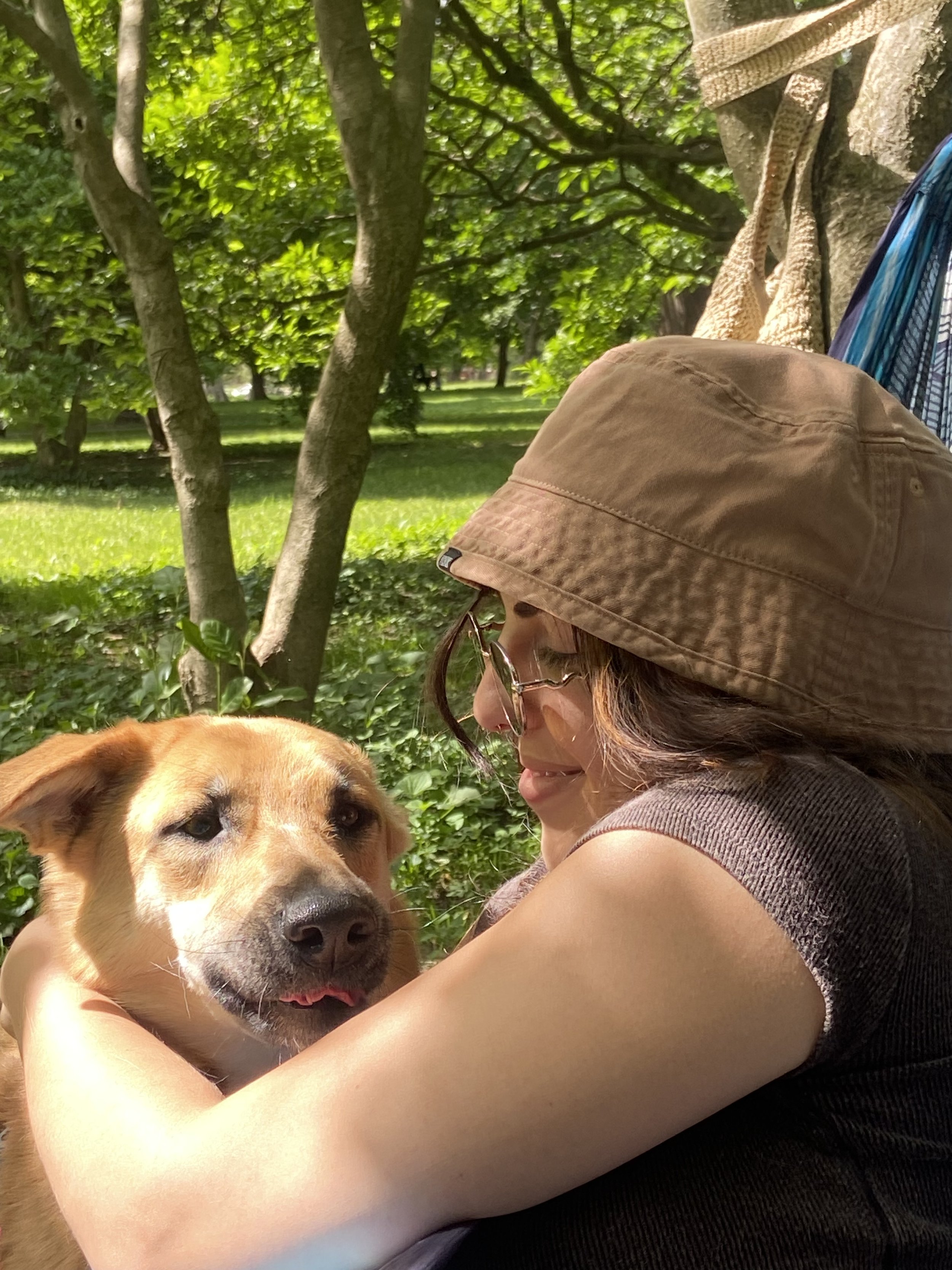 A woman wearing glasses and a brown sun hat is hugging a tan dog with a black nose in a park with green grass and trees.