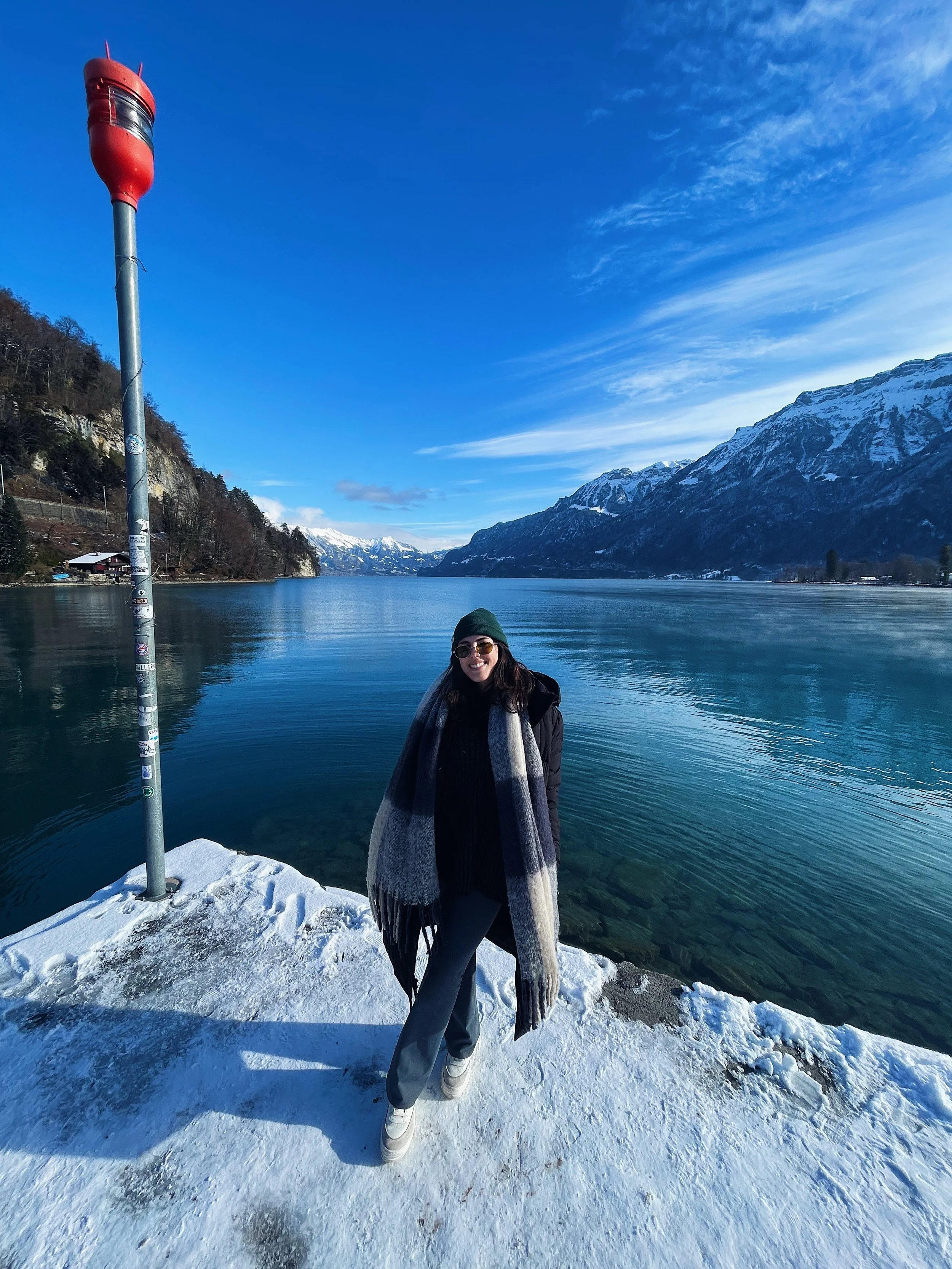 A woman standing on snow-covered ground by a lake, wearing a green beanie, sunglasses, a black coat, a large gray and black scarf, and white boots, with snow-capped mountains and a clear blue sky in the background.