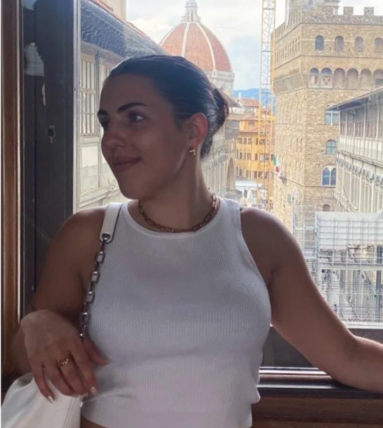 A woman with dark hair tied back, wearing a white sleeveless top, sitting by a window with a cityscape background, including historic buildings and a dome, in Florence, Italy.