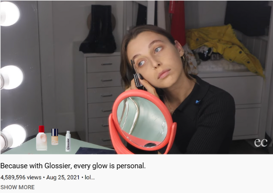 A woman at a vanity mirror applying makeup, with beauty products on the table, in a bedroom.
