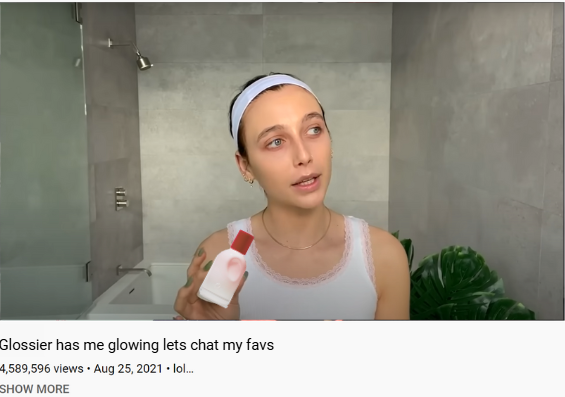 A woman in a bathroom holding a bottle of skincare product, wearing a white tank top and a headband.