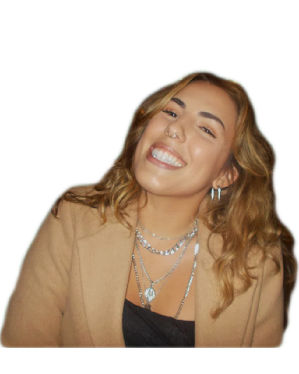 A woman with curly blonde hair smiling, wearing layered silver necklaces, hoop earrings, a nose ring, and a tan blazer over a black top.