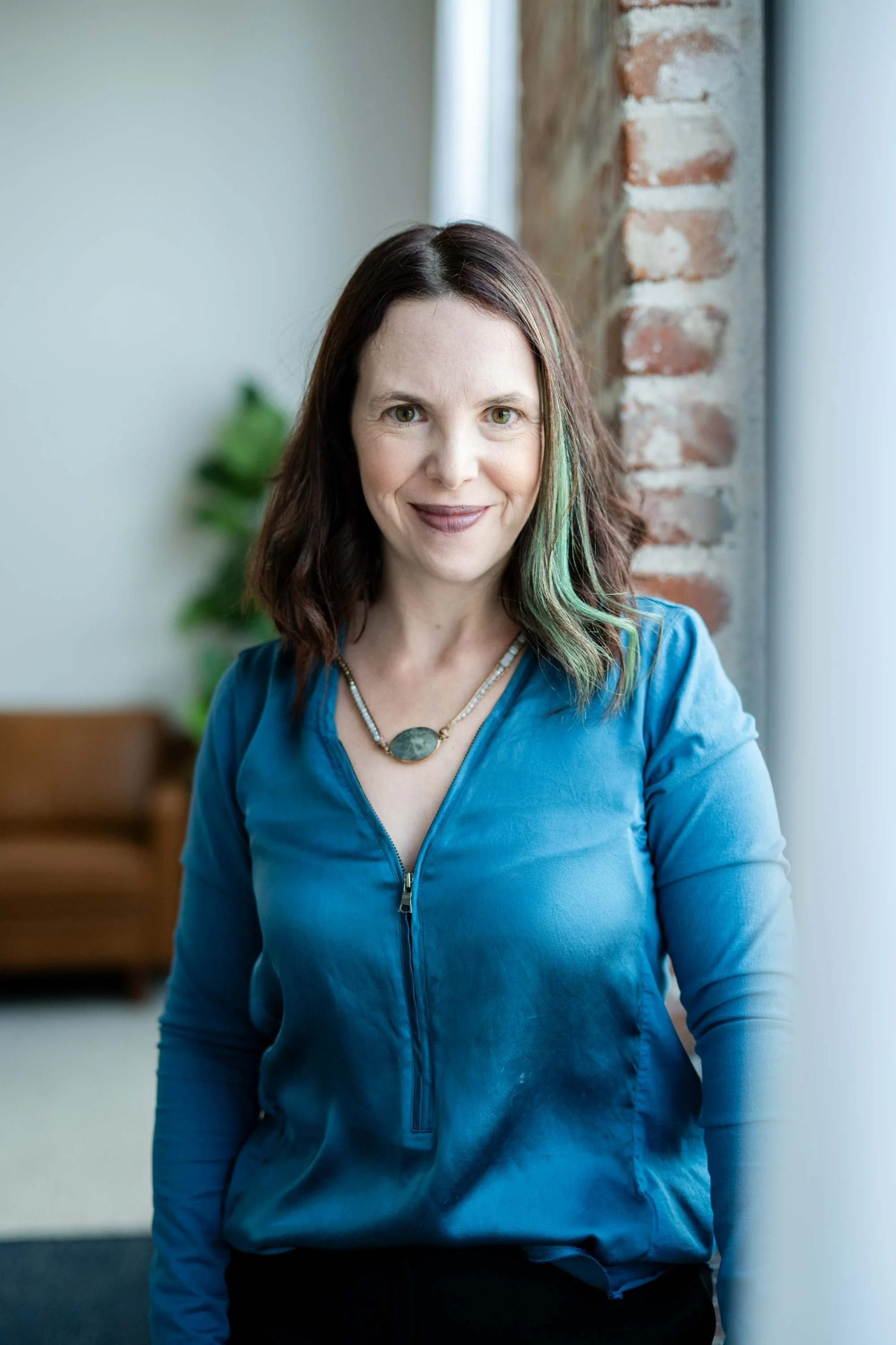 A woman with shoulder-length dark hair with green streaks, wearing a blue zip-up top and a statement necklace, standing indoors near a brick wall and smiling at the camera.