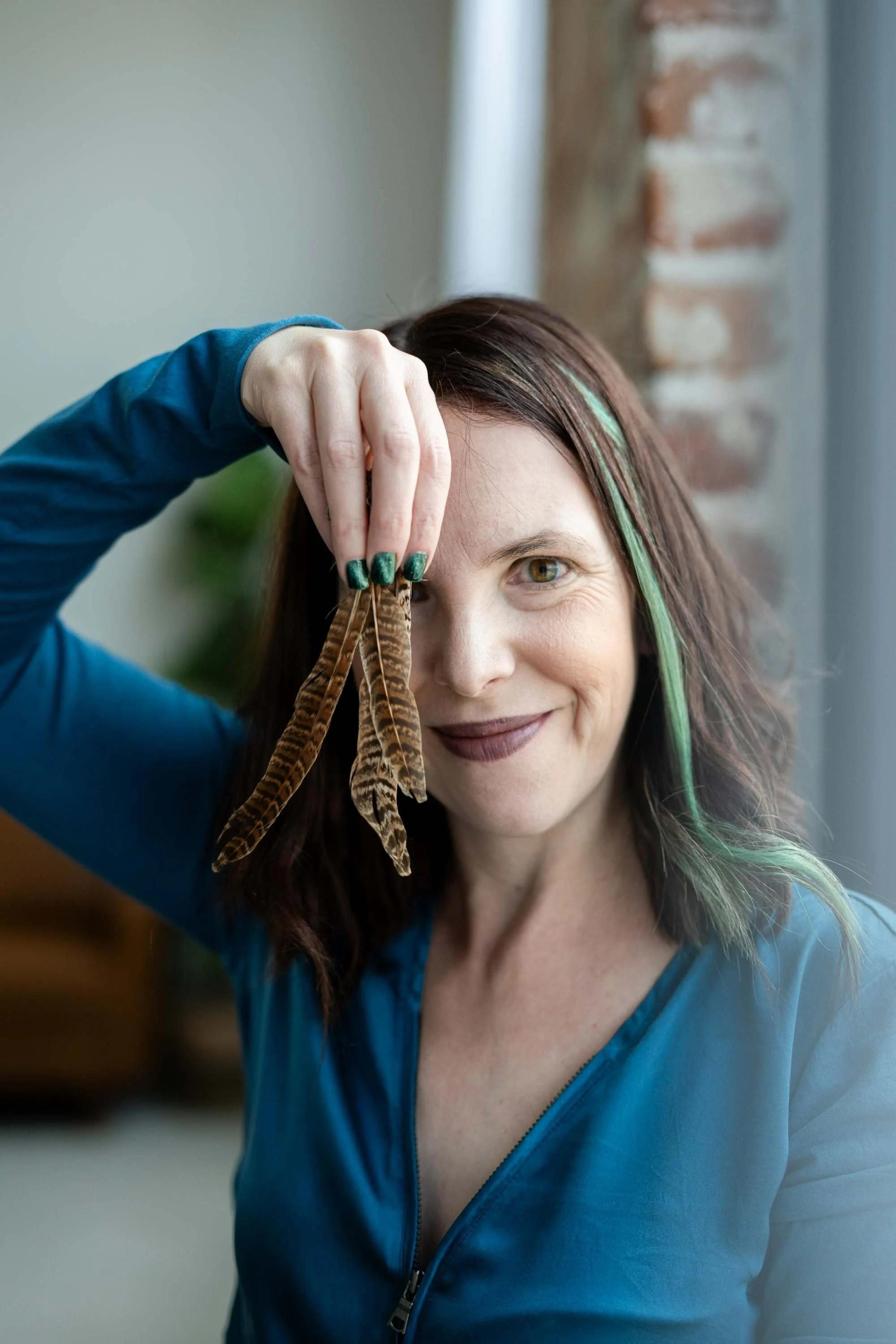 Woman with dark hair and green highlights holding a feather near her eye, smiling