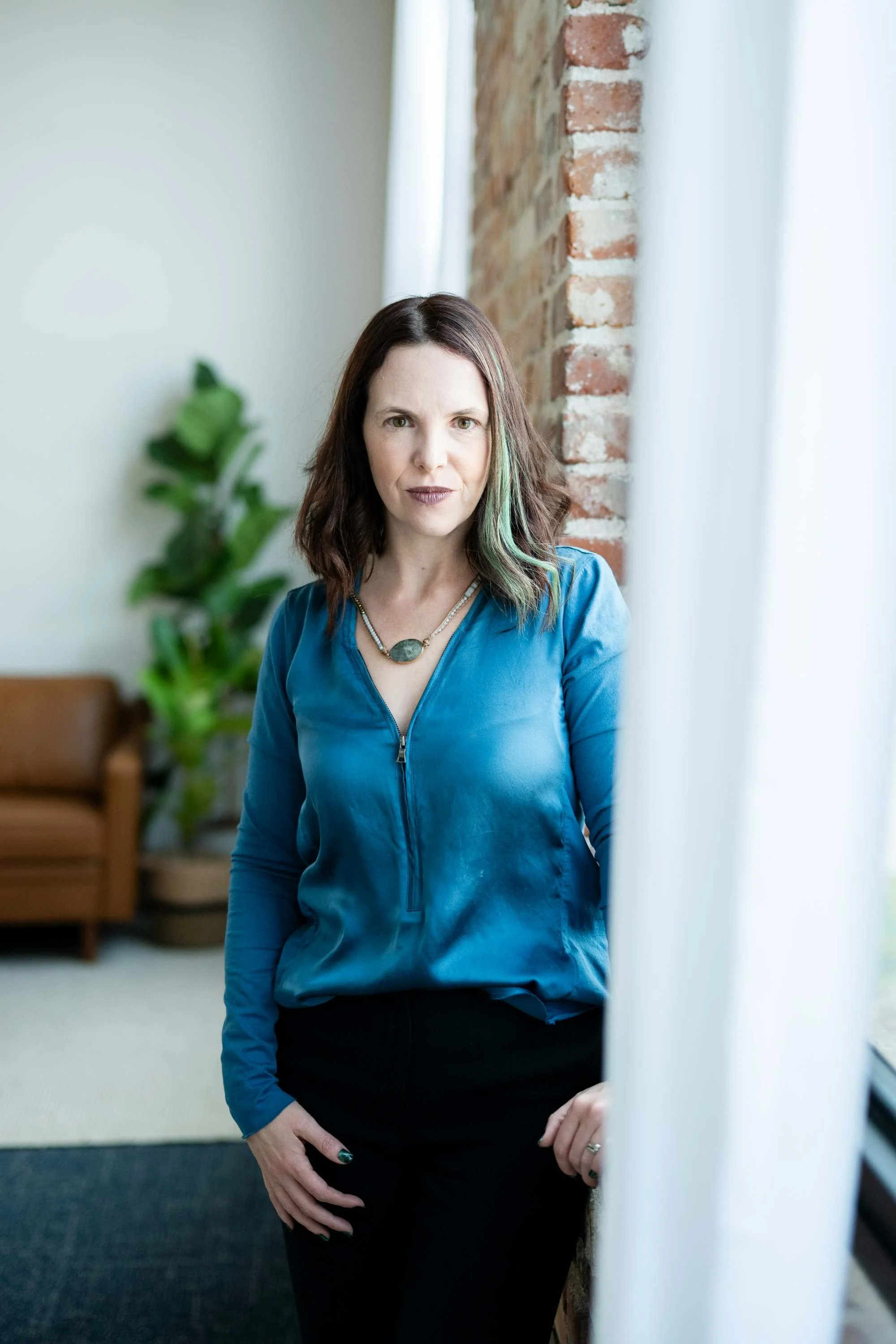 A woman with shoulder-length brown hair and fair skin standing indoors near a brick wall and window, wearing a blue satin blouse, black pants, and a necklace.
