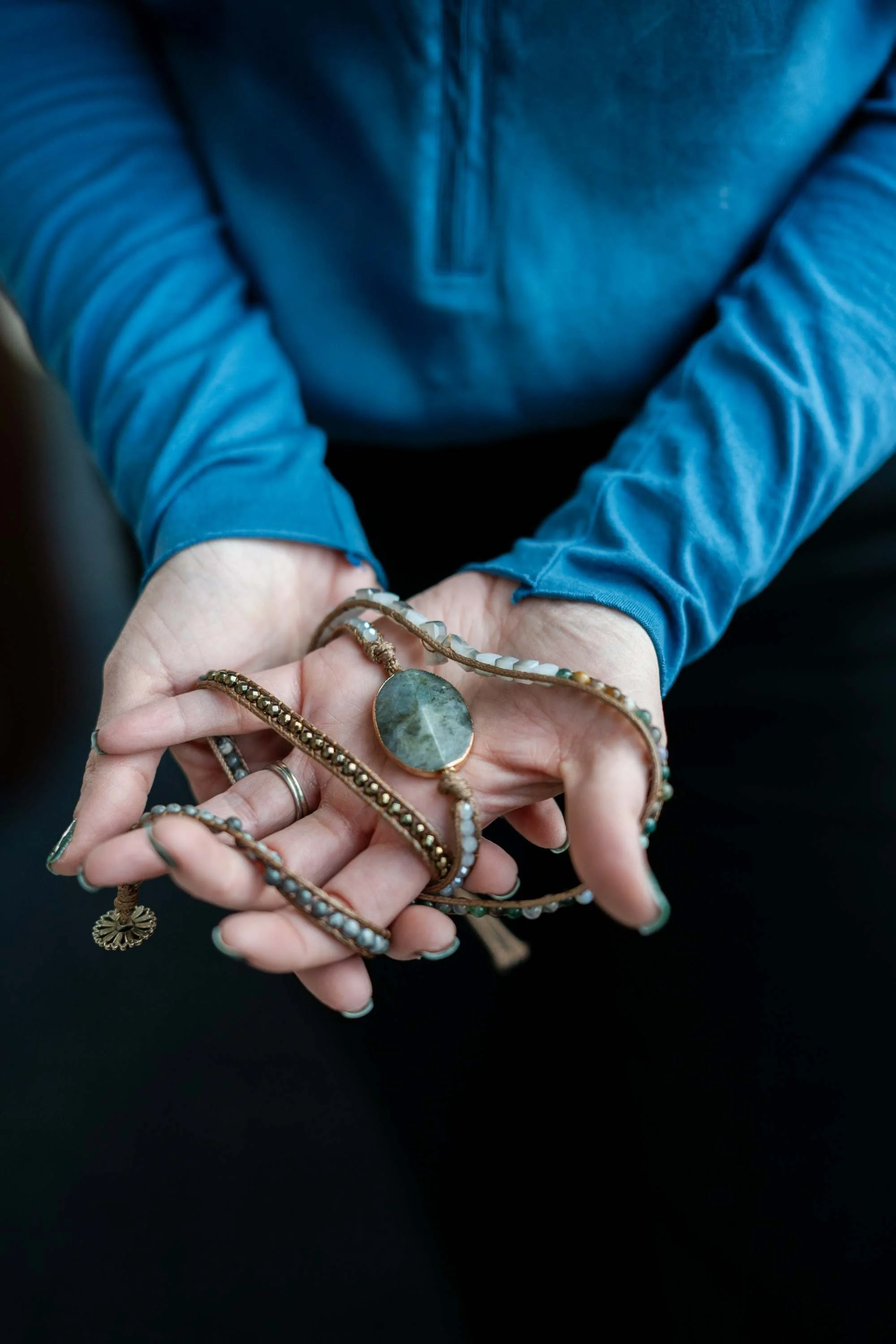 Person wearing a blue long-sleeve shirt holding several pieces of jewelry, including rings and a necklace, with a blurred background.