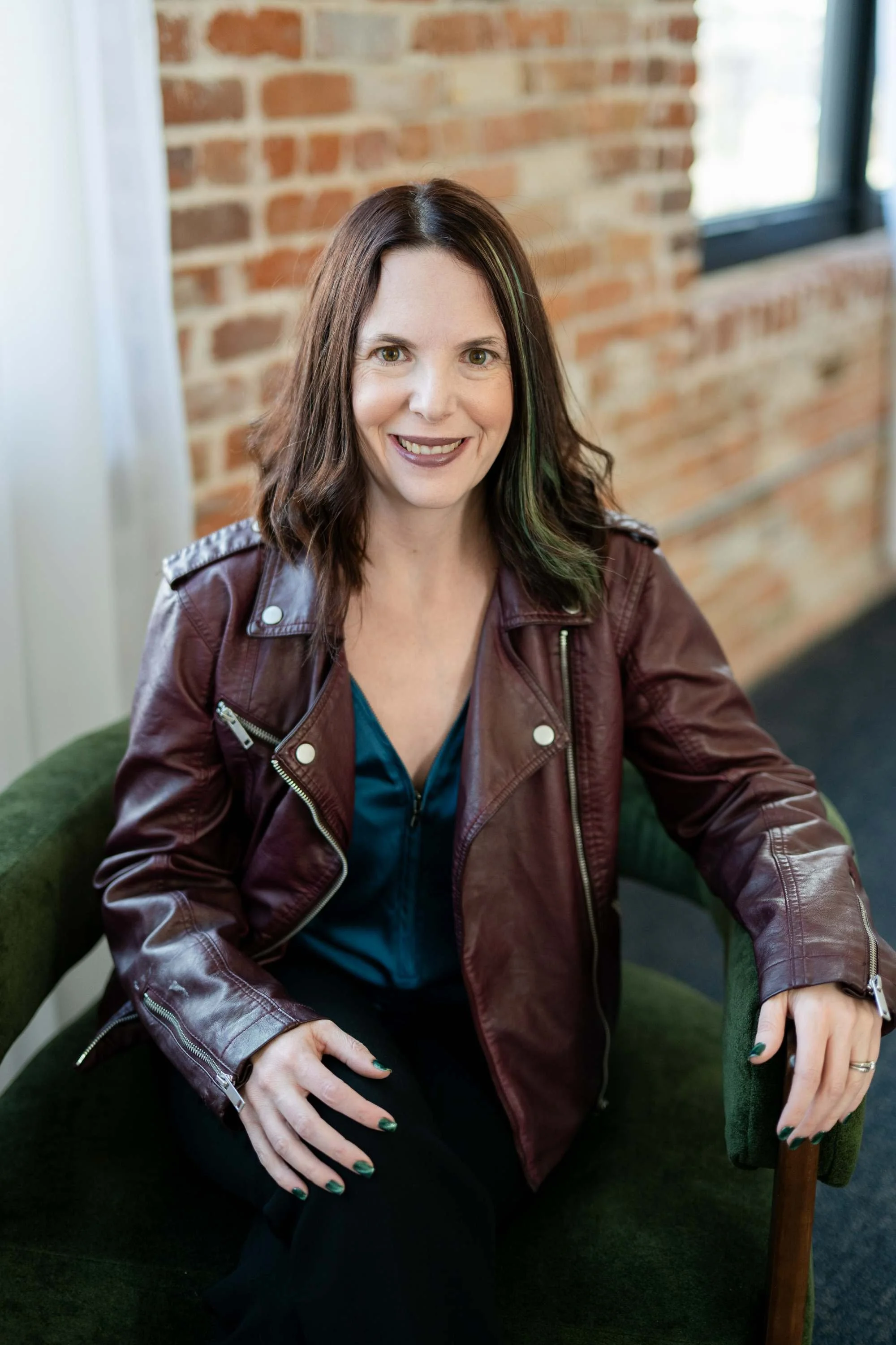 A woman with shoulder-length brown hair, wearing a maroon leather jacket and a blue top, sitting on a green chair in a room with a brick wall and a window.