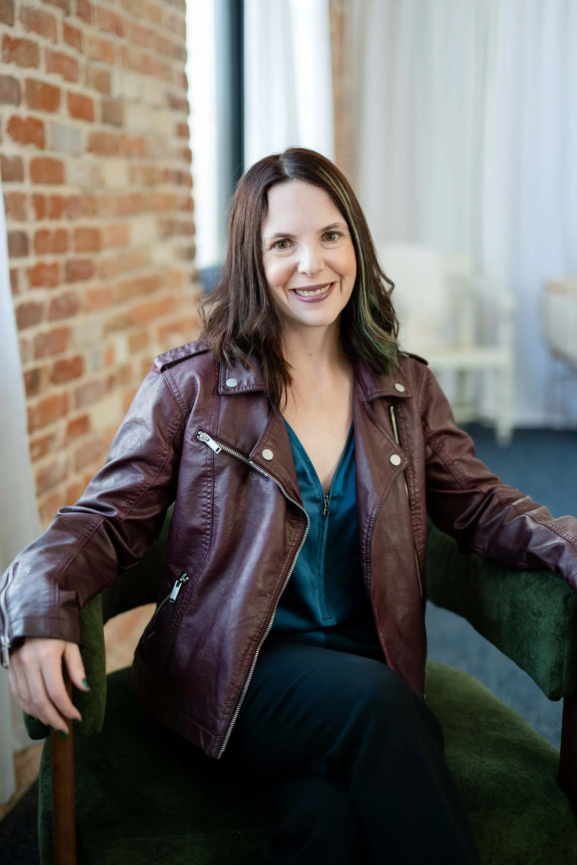 A smiling woman with shoulder-length dark hair and green highlights, wearing a maroon leather jacket over a teal blouse, sitting on a green chair in a room with a brick wall and large window with white curtains.