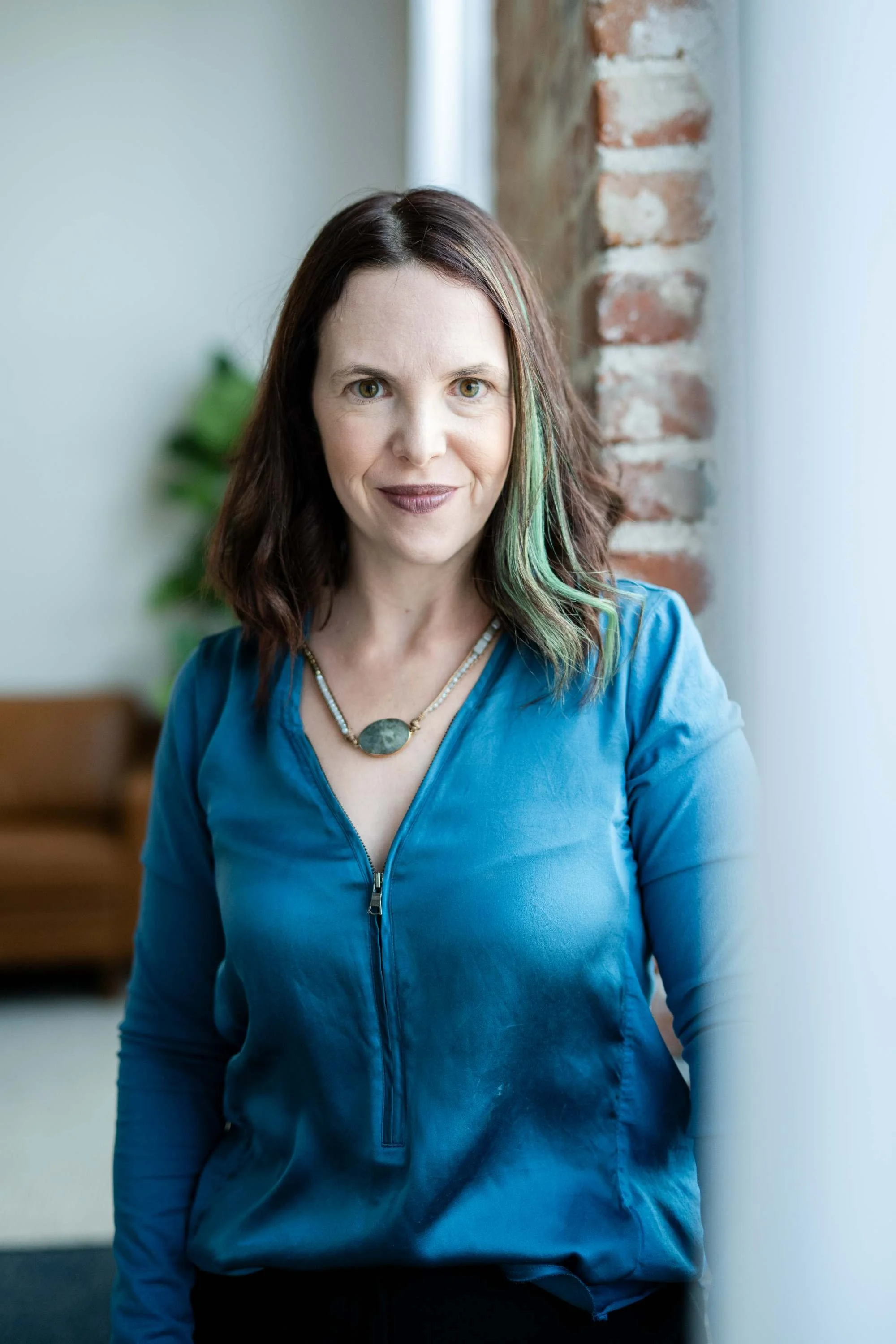 A woman with shoulder-length dark hair and a hint of green highlights, wearing a blue zip-up top and a necklace, standing beside a window with a brick wall and a blurred couch in the background.