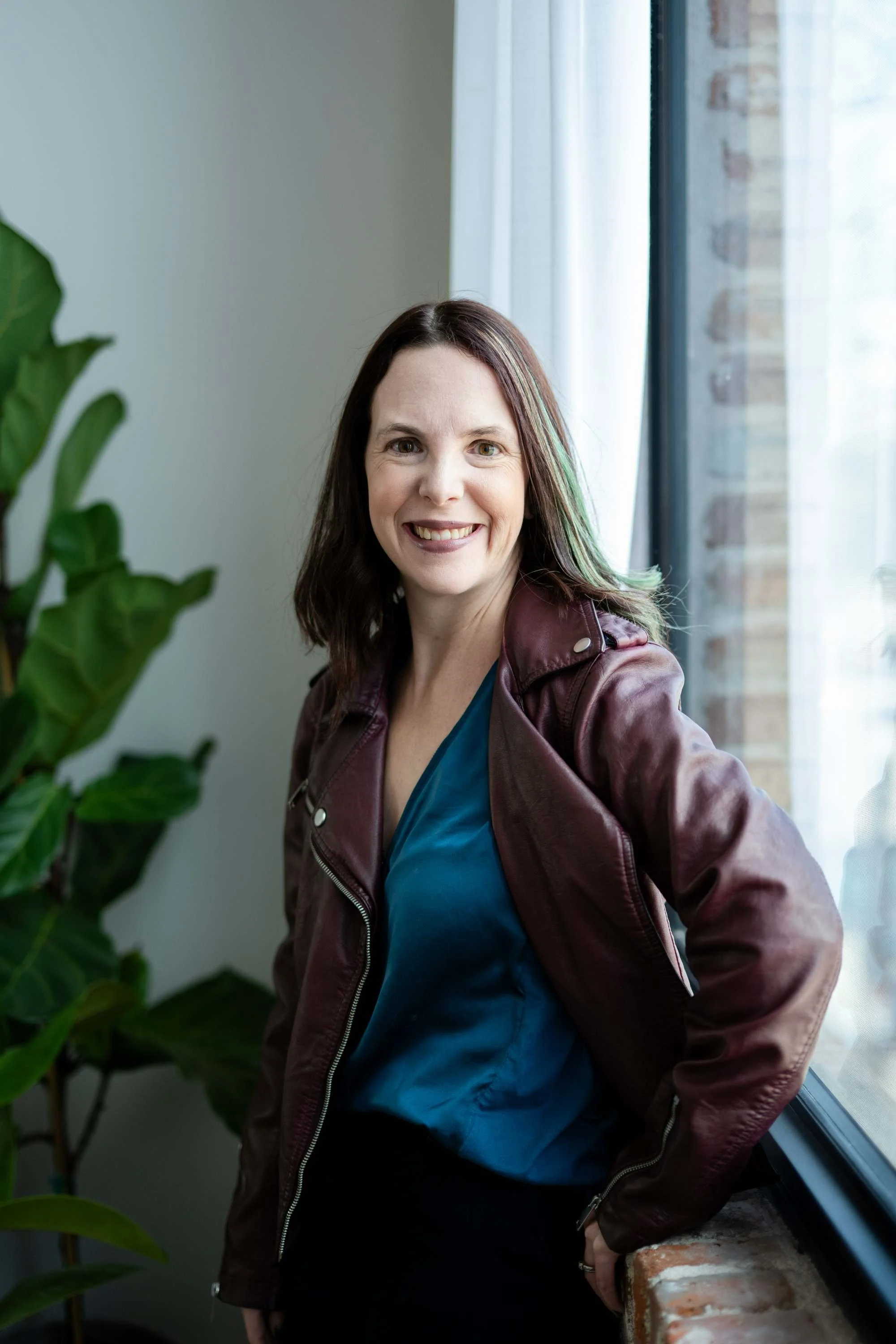 Smiling woman with dark brown hair wearing a maroon leather jacket and a teal blouse, standing next to a window with a brick wall outside, with large green plants nearby.