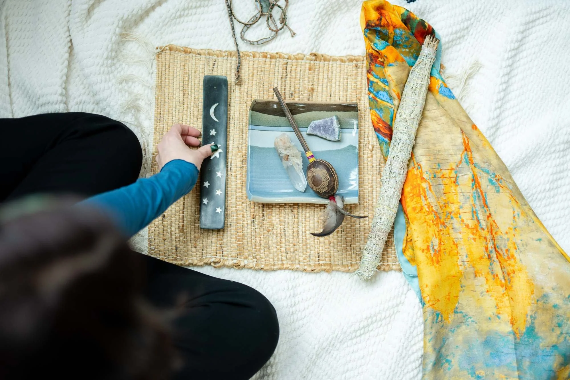 A person sitting on a woven mat with various objects used for a spiritual or ritual practice: a black rectangular object with white moon and star designs, a square dish with rocks and a shell, a spoon, a rolled-up colorful cloth or curtain, and a narrow black and rainbow-colored object. The person's hand is reaching towards the black object.