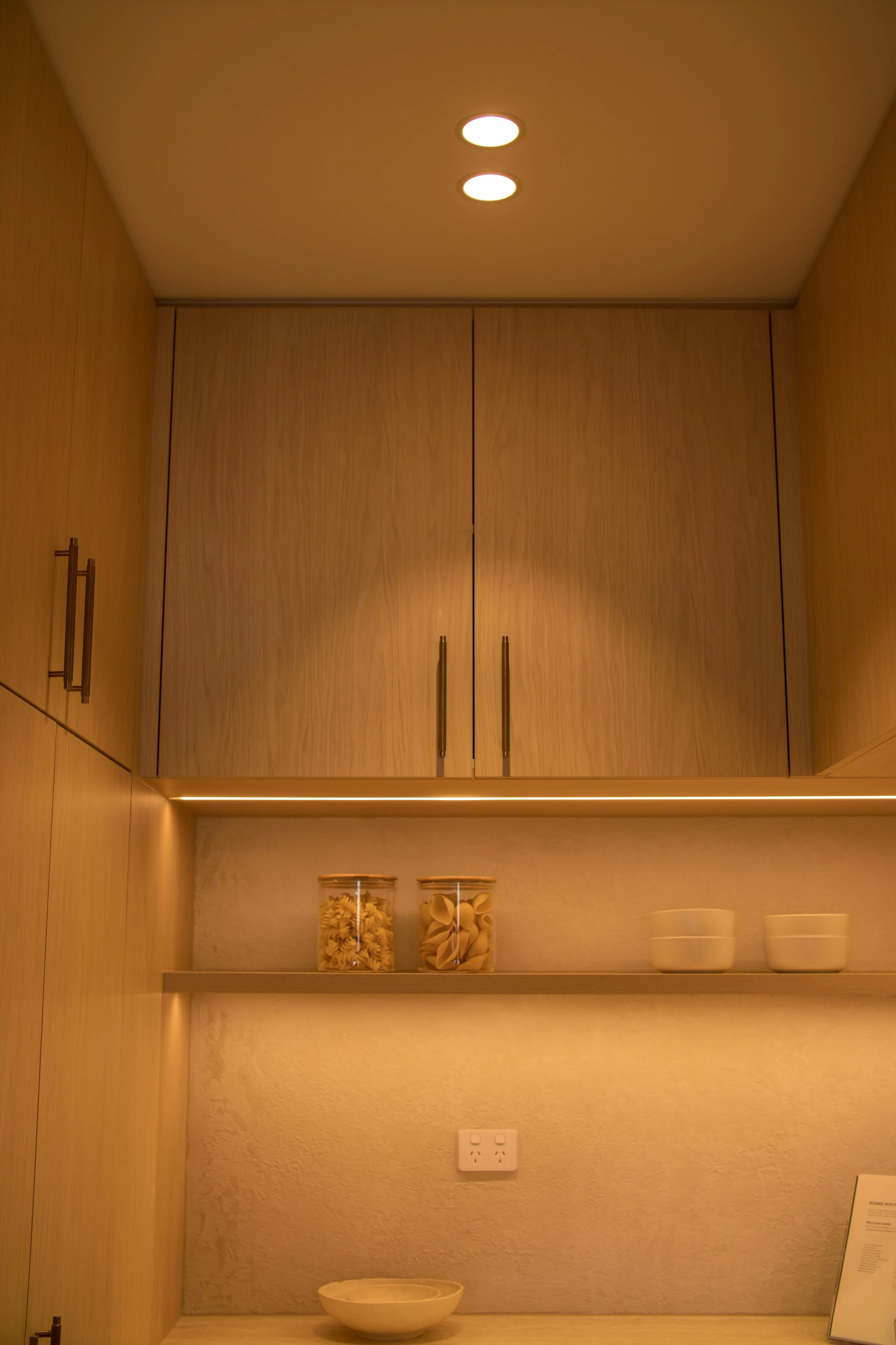 Kitchen cabinets with two glass jars of pasta and two white bowls on a shelf, with a power outlet and a bowl on the countertop below.