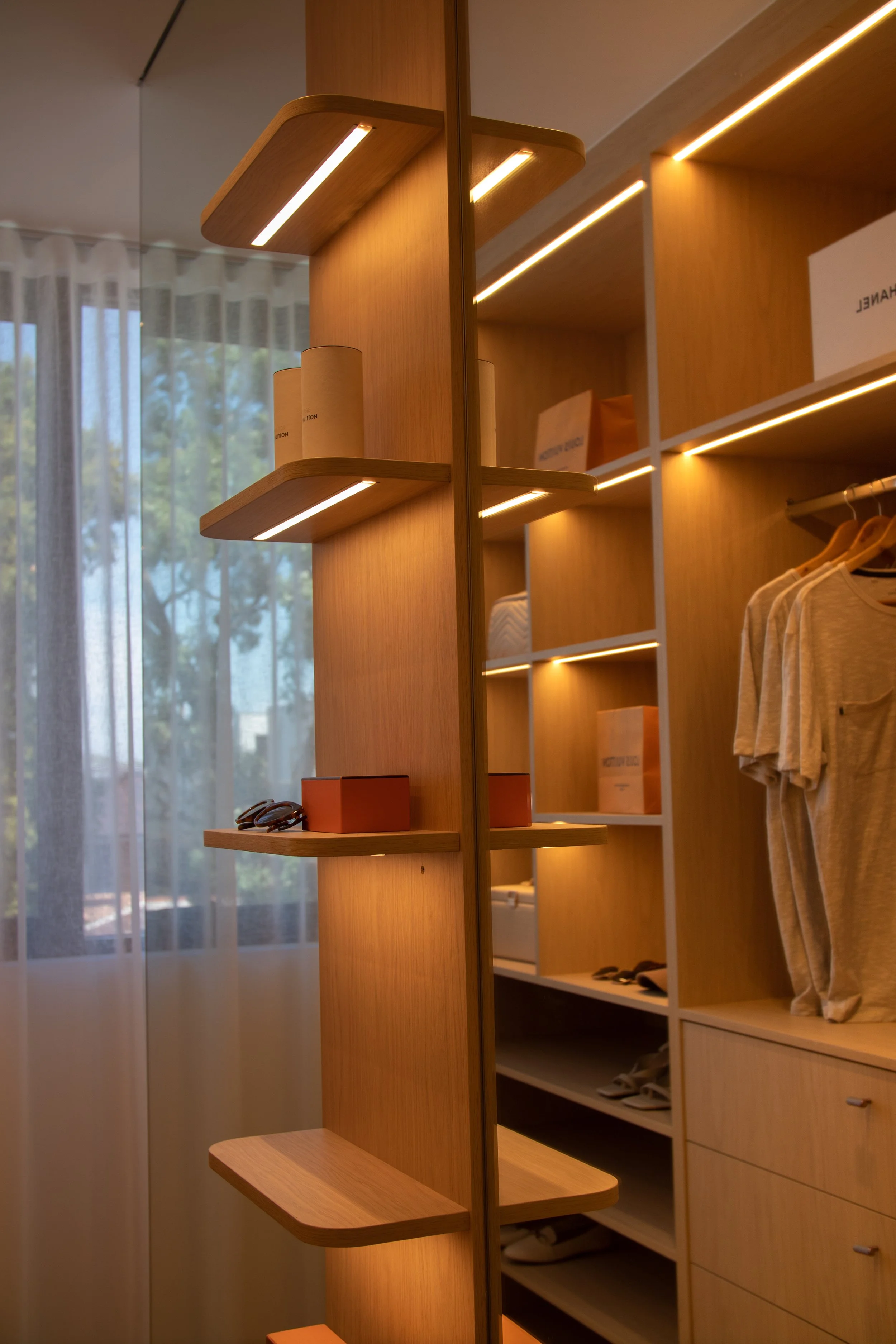 Wooden closet with shelves and drawers, inside a room with a window and sheer curtains, showing clothing, boxes, and accessories.