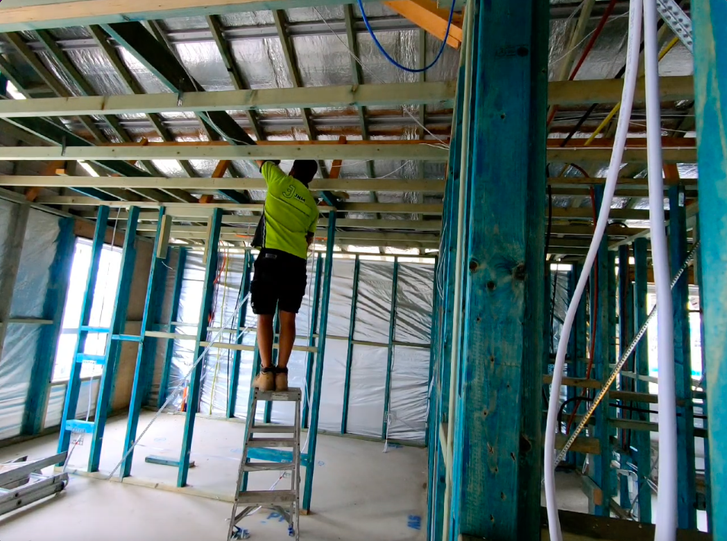 A construction worker in a bright yellow shirt and black shorts standing on a small ladder, working on the ceiling framework inside a building under construction. The room has exposed framing, electrical wiring, and plastic sheeting covering some areas.