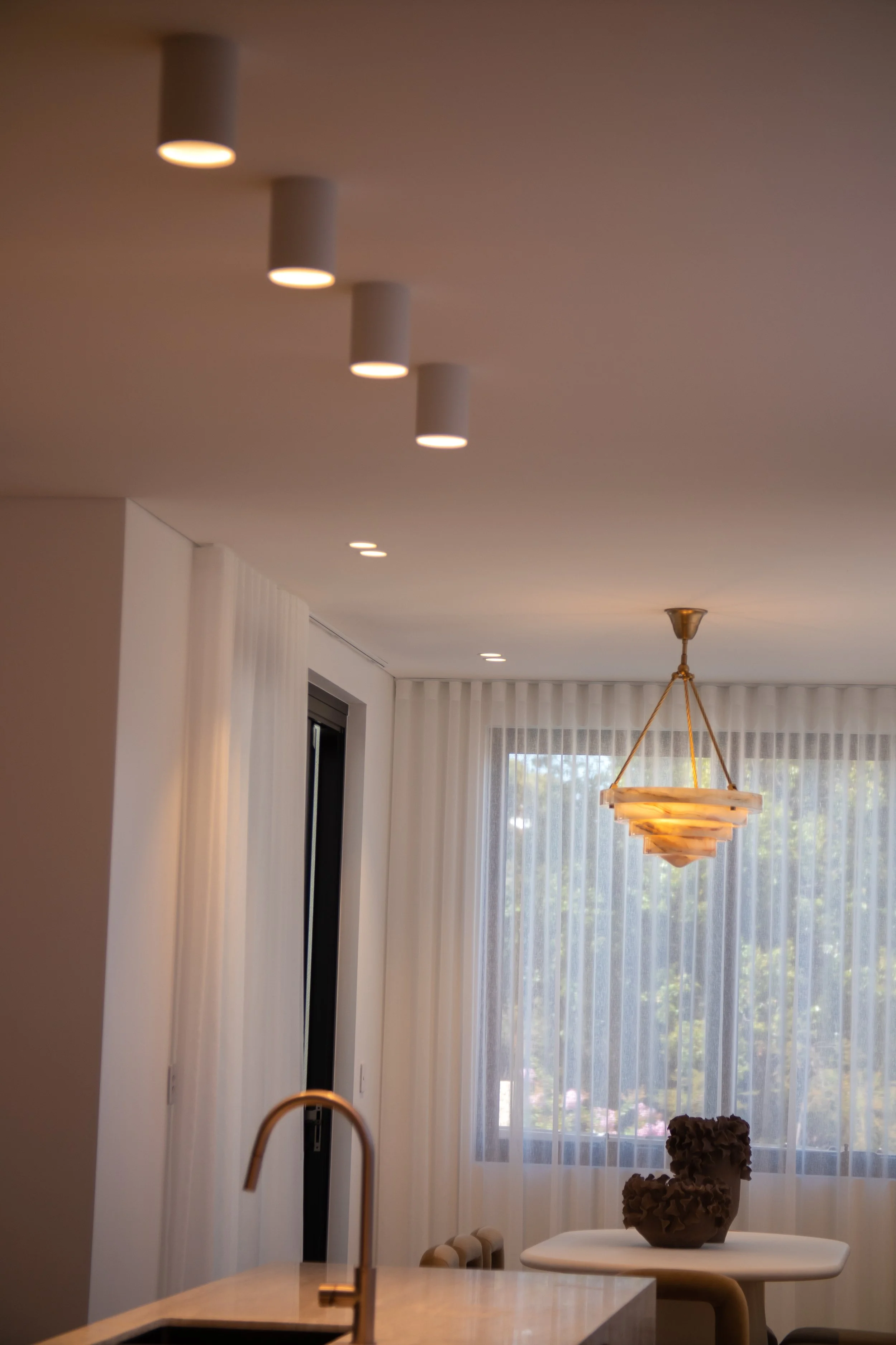 Interior view of a modern living space with a ceiling featuring multiple recessed lights, a brass chandelier, sheer white curtains, a small round table with a decorative brown sculpture, a window with trees outside, and part of a kitchen island with a gold-colored faucet.
