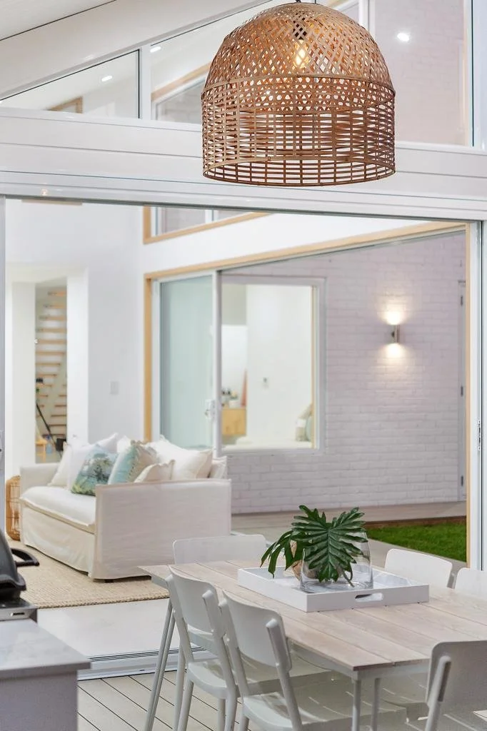 Contemporary dining area with a wooden table, white chairs, and a large green plant in a glass vase. Lounge area with a white sofa and decorative pillows, and a woven rattan pendant light hanging from the ceiling. Large sliding glass doors open to an outdoor space with brick walls and wall-mounted lighting.