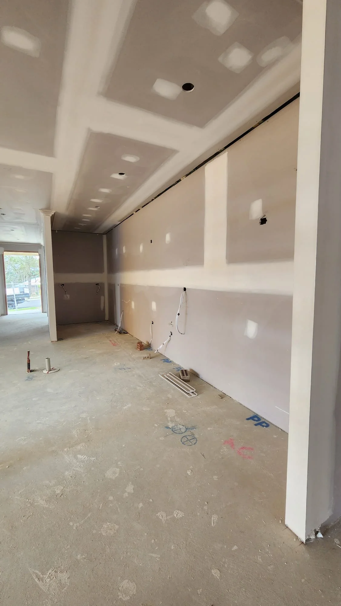 Interior of a room under construction with drywall and a ceiling with recessed lighting holes, electrical wiring visible, and an unfinished floor.