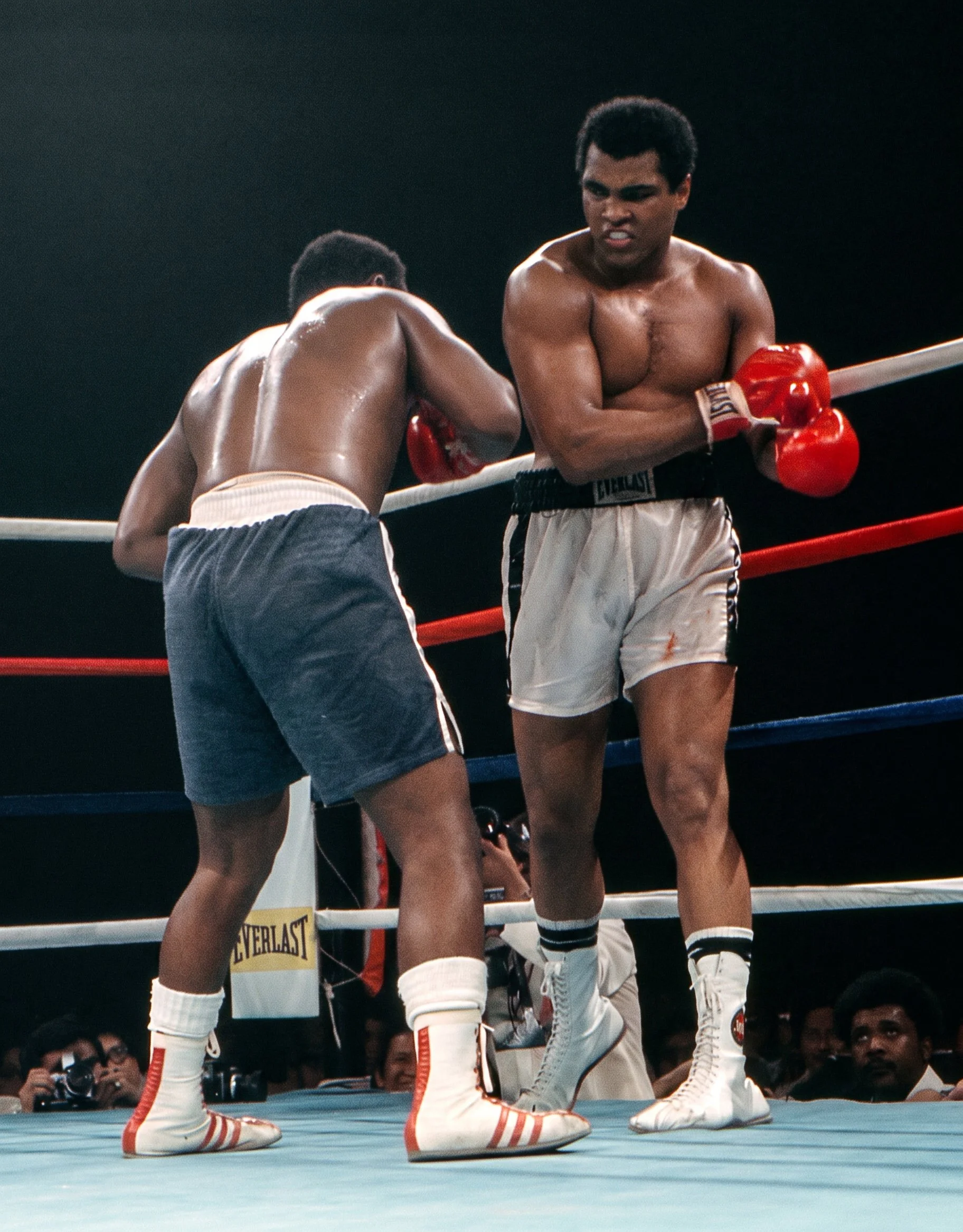 An exhausted Joe Frazier doubles over in front of Muhammad Ali during the later rounds of Ali-Frazier III, with blood visible on Ali’s white trunks.