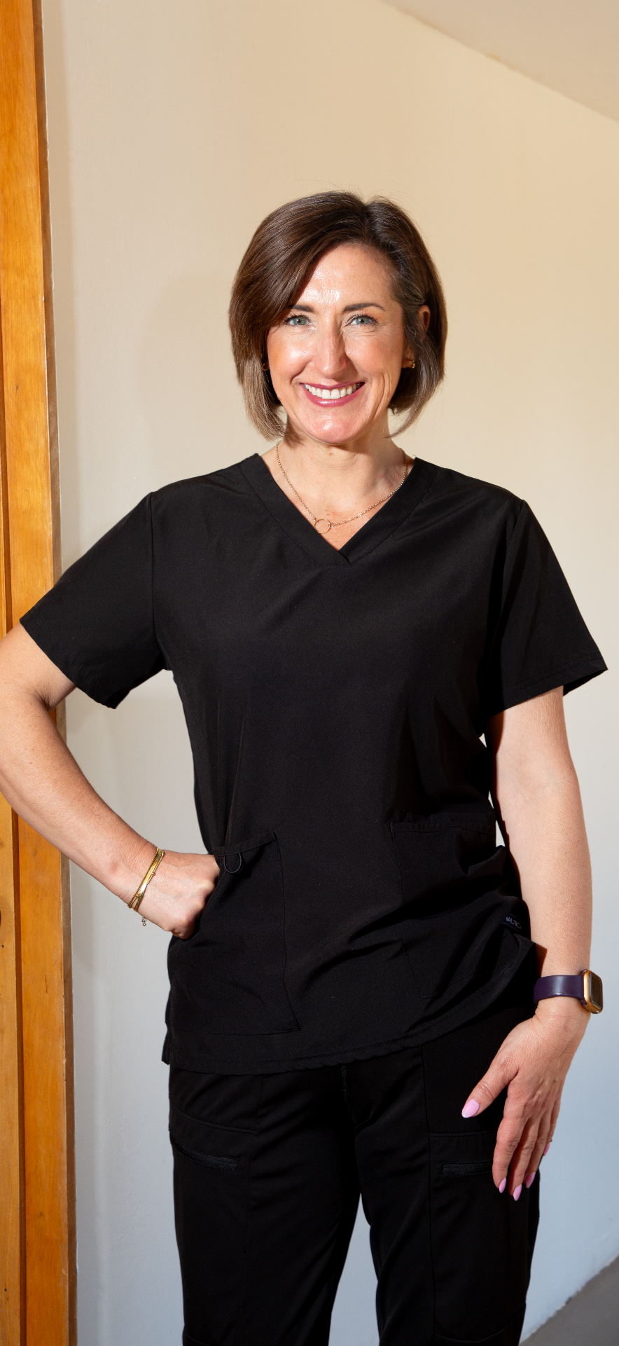 A woman wearing black medical scrubs, smiling and standing with her hand on her hip in a room with a wooden door and beige wall.