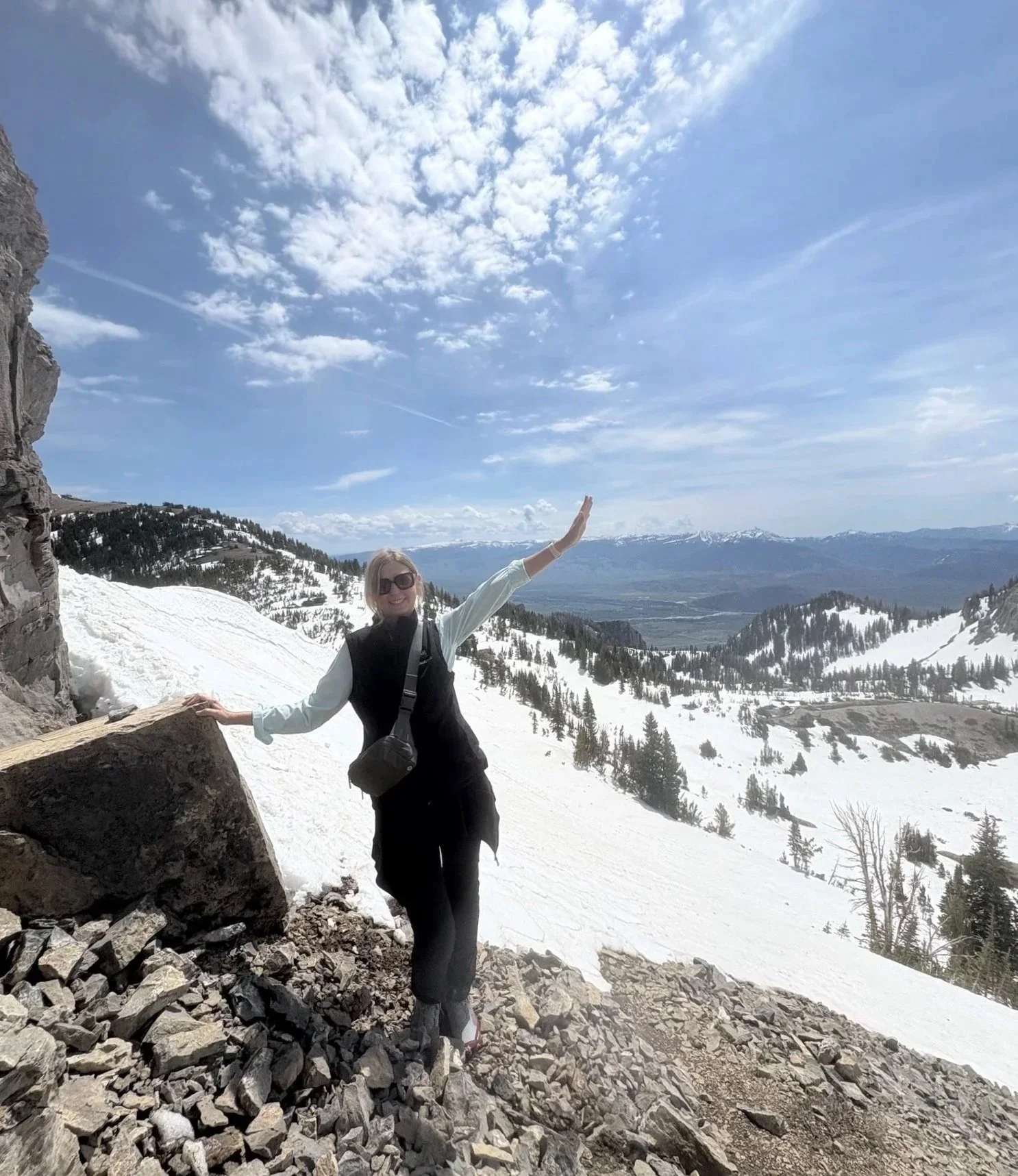 Woman standing on a rocky mountain trail surrounded by snow-covered peaks.