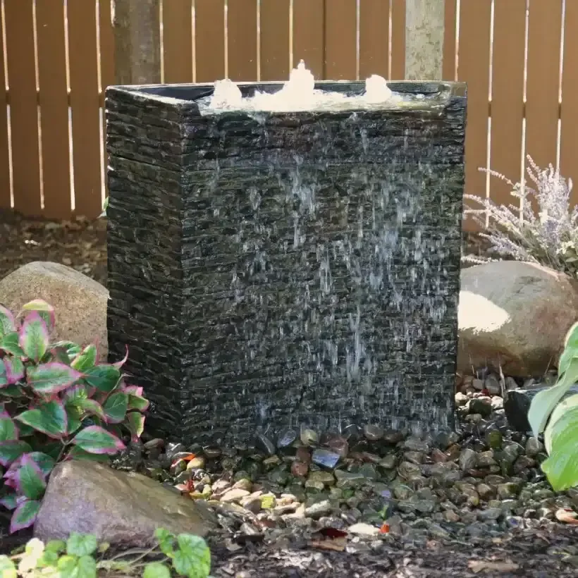 A water feature with water cascading down a dark, textured stone cubic structure, surrounded by rocks and greenery in a garden setting.
