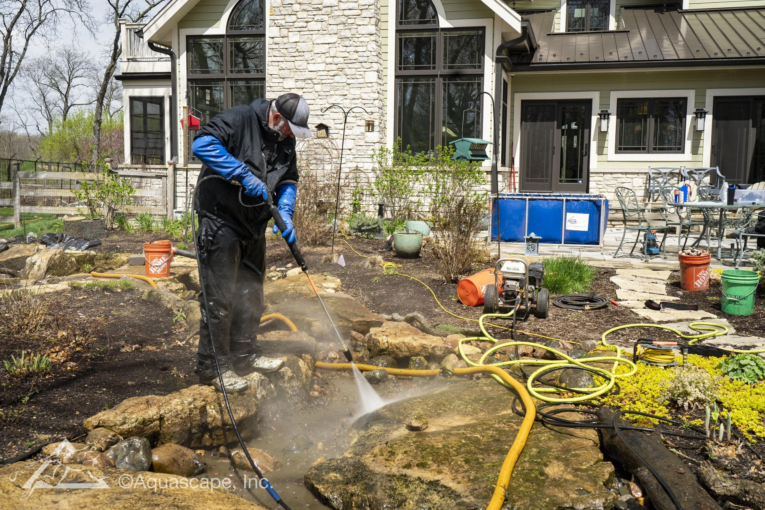 Professional pond cleaning service with technician performing a spring pond cleanout and pressure washing rockwork around a backyard water feature.