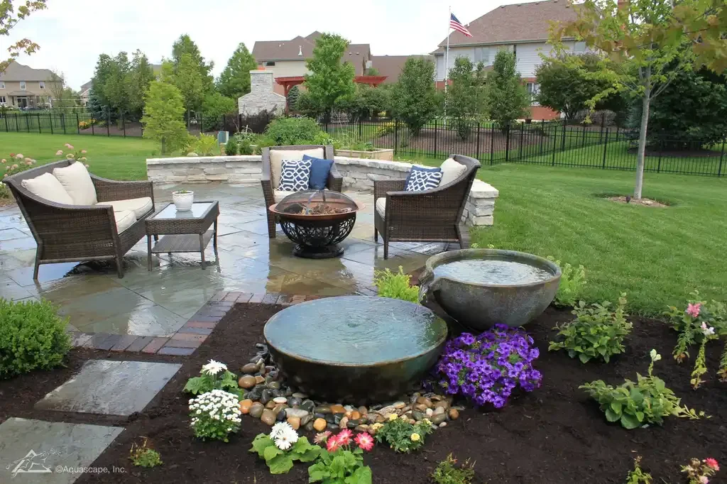 A backyard patio with outdoor seating, including wicker chairs with cushions and pillows, a small side table, and a fire pit. In the foreground, there are two large planters with water and colorful flowering plants nearby. The yard is enclosed by a black fence, with trees, shrubs, and houses visible in the background.