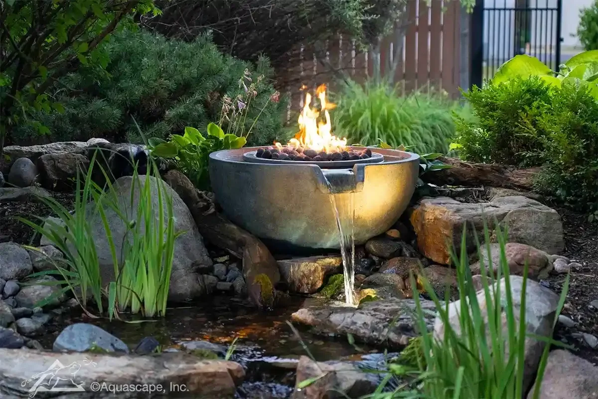A backyard garden with a small fountain featuring a fire sculpture in a round basin, surrounded by rocks and lush green plants.