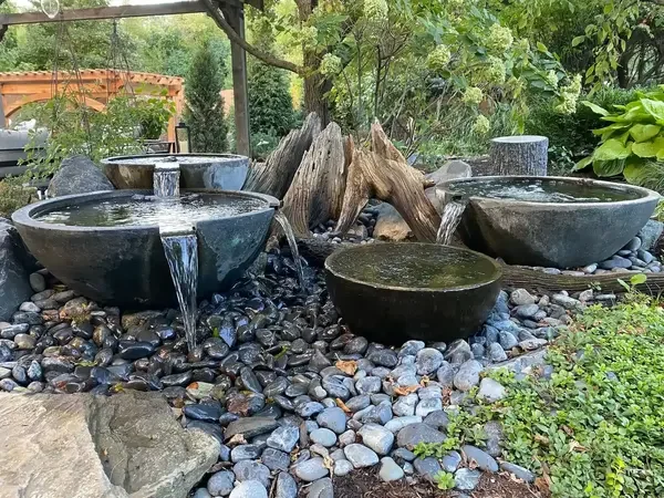 Three stone water bowls of different sizes with water flowing from one bowl to another, set among smooth black and gray river stones in an outdoor garden with trees and plants.