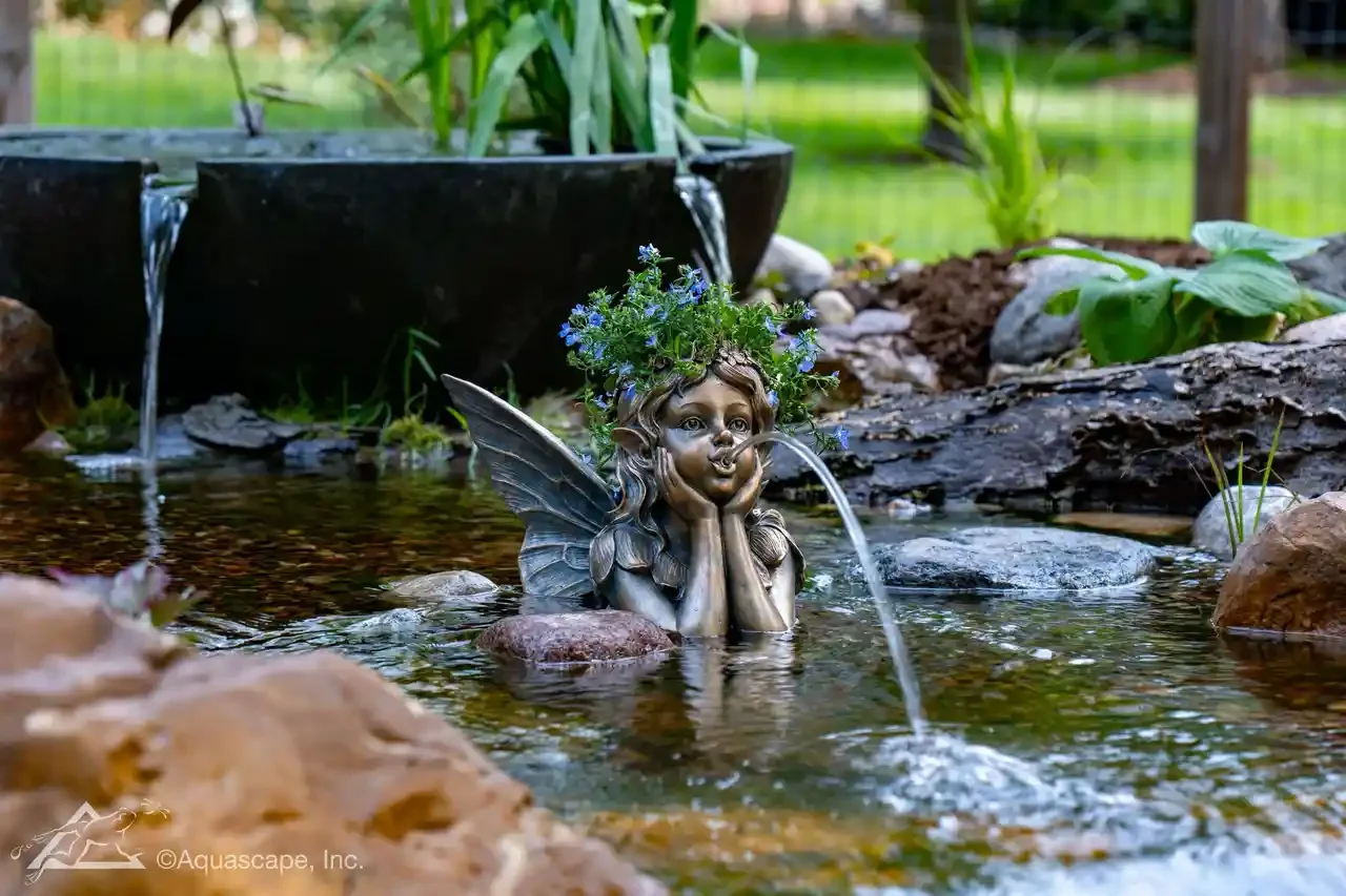 A garden fountain featuring a fairy statue with wings, surrounded by rocks and plants, with water flowing from the fairy's nose and a pot with water in the background.
