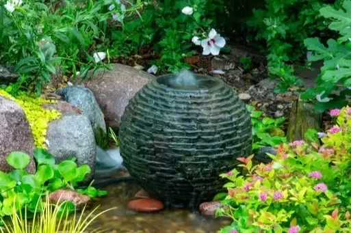 A black spherical garden fountain with water flowing from the top, surrounded by various green plants and colorful flowers in a lush garden setting.