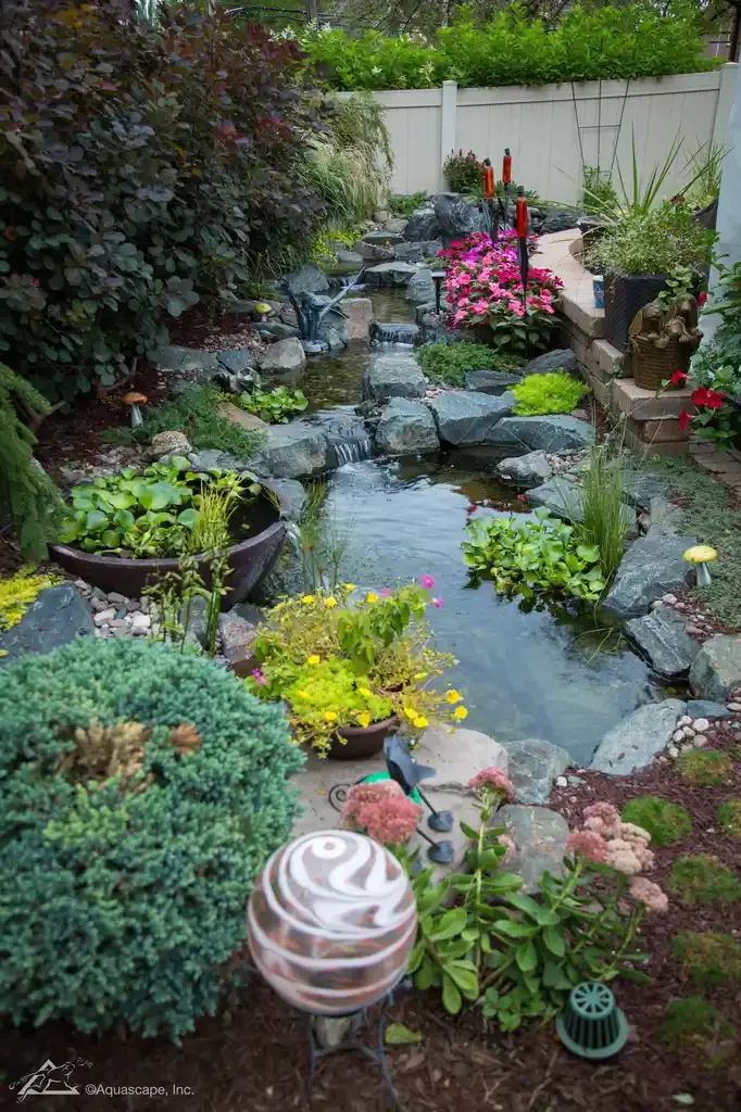 A backyard with a small, rocky pond, surrounded by various blooming plants and shrubs. There's a white fence in the background, and a raised brick platform with potted flowers. Water features and garden decorations are also visible.