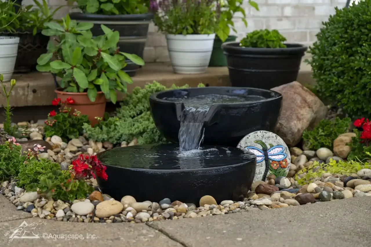 A small garden with a black stone water fountain, surrounded by colorful flowers and potted plants on a paved surface.