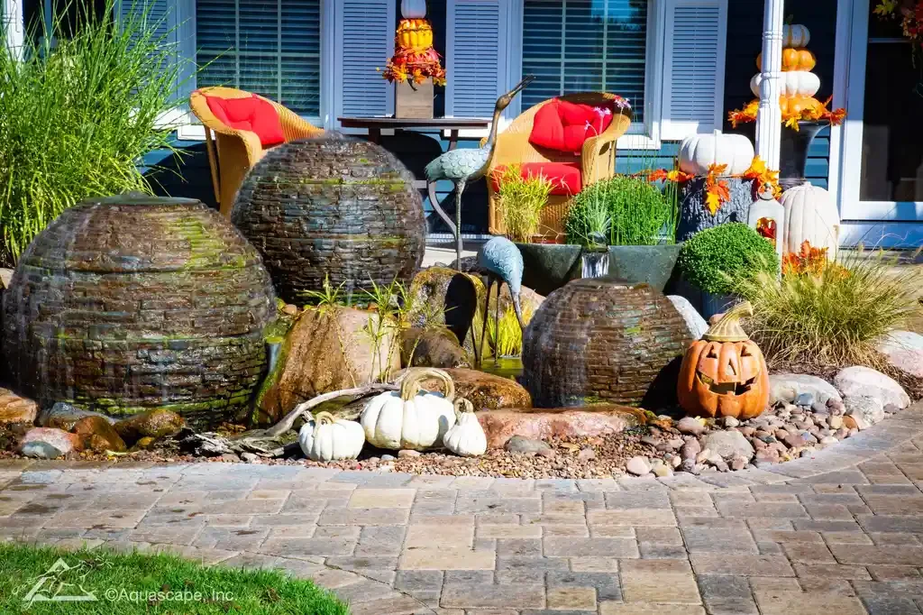 Decorative backyard garden with water fountain feature, white and orange pumpkins, a ceramic stork statue, lush green plants, and outdoor furniture with red cushions, all set on a paved patio in front of a house.
