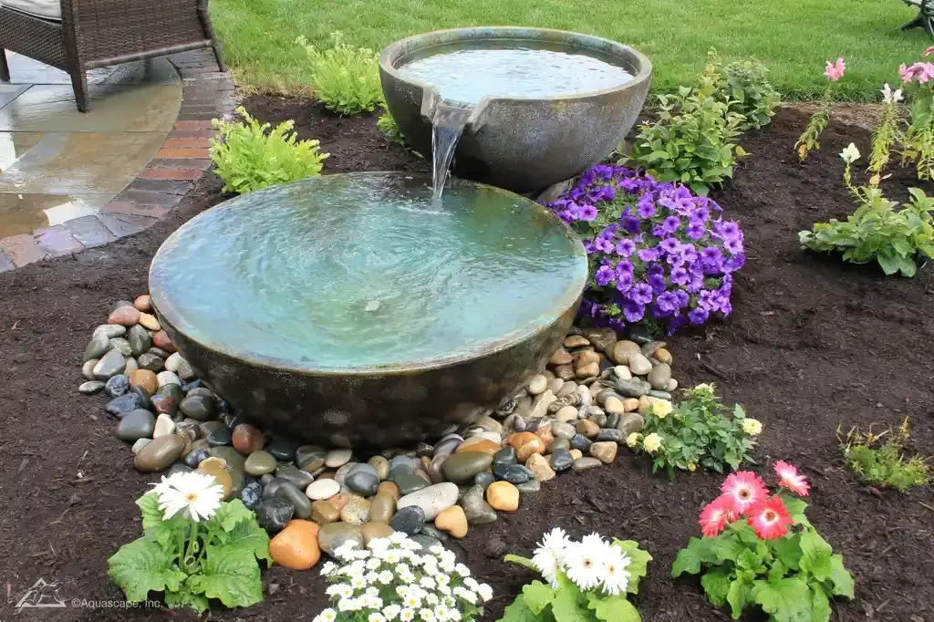 Two-tier stone water fountain surrounded by colorful flowers and gravel in a garden.