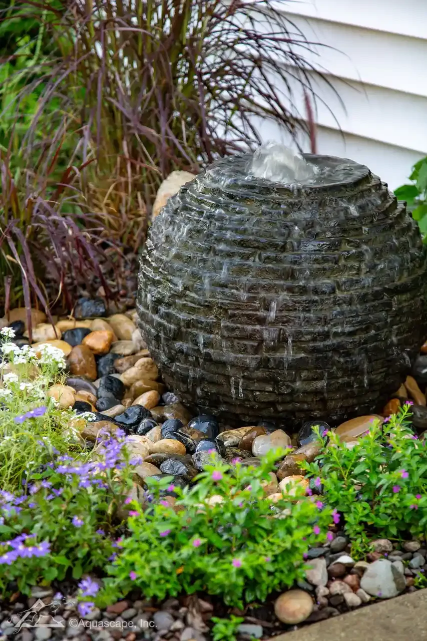 A black, round, textured garden water fountain with water flowing from the top, surrounded by small multicolored pebbles and flowering plants with purple and white blooms.