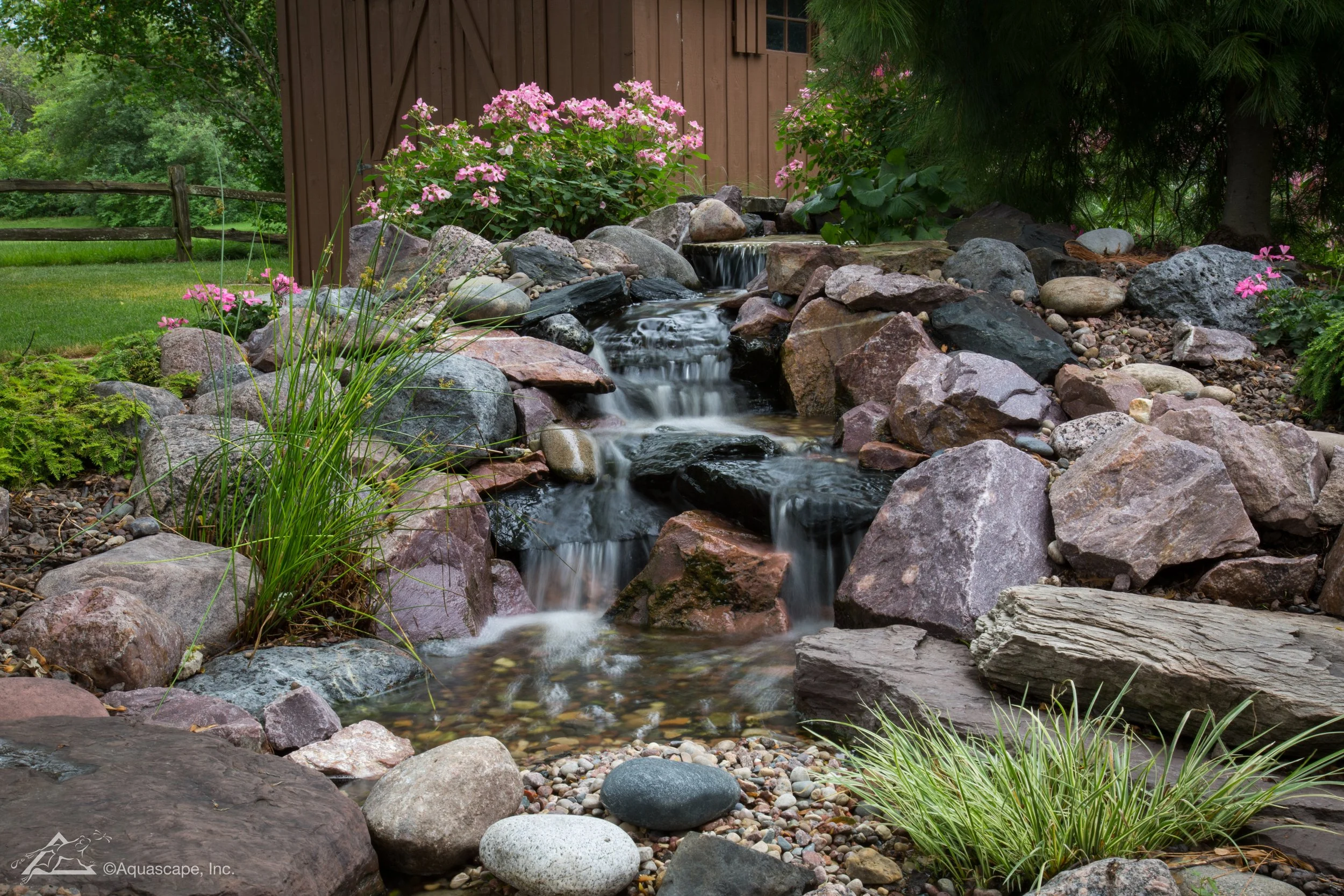 Aquascape-style pondless waterfall with natural boulders, cascading water, and colorful garden plantings in a residential landscape.