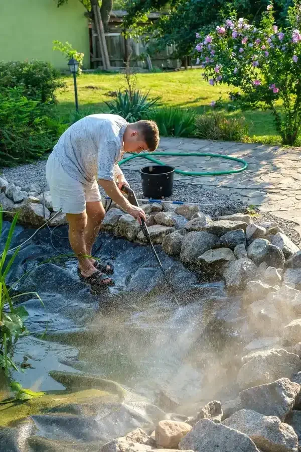 A person is cleaning a small pond in a garden with rocks around it, using a net or a tool, while water is being filtered or disturbed.