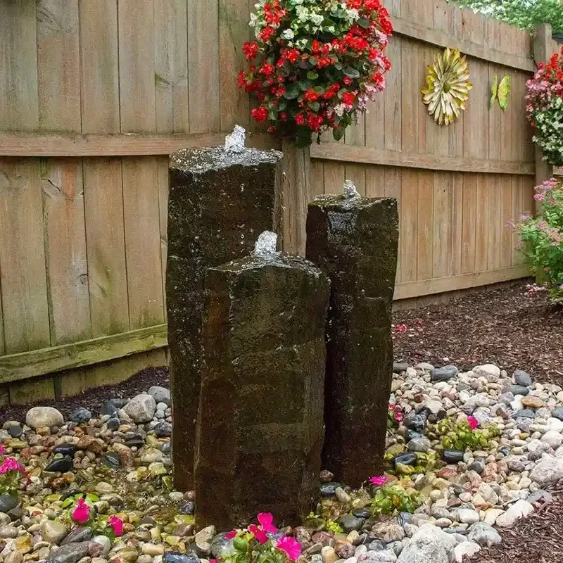 Three dark upright stones with water flowing over them, arranged in a garden setting with colorful flowers, small rocks, and a wooden fence in the background.