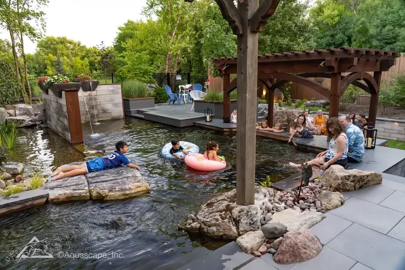 A backyard pool area with children swimming and playing in the water, adults sitting on the rocks and under a pergola, and patio furniture in the background.