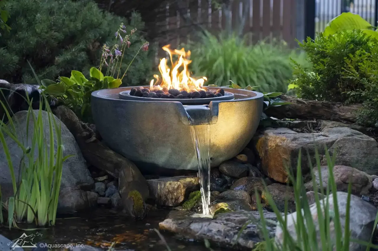 Decorative outdoor water feature with a small waterfall flowing into a pond, surrounded by rocks, plants, and greenery.