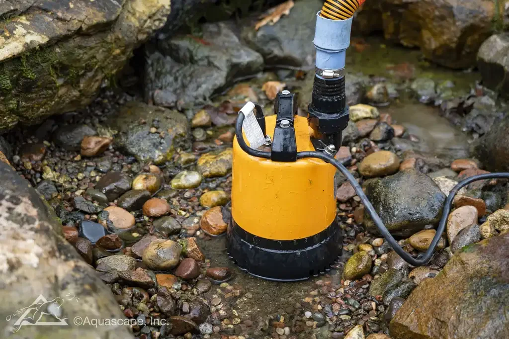 A yellow submersible water pump placed in a rocky shallow stream surrounded by rocks and small pebbles.