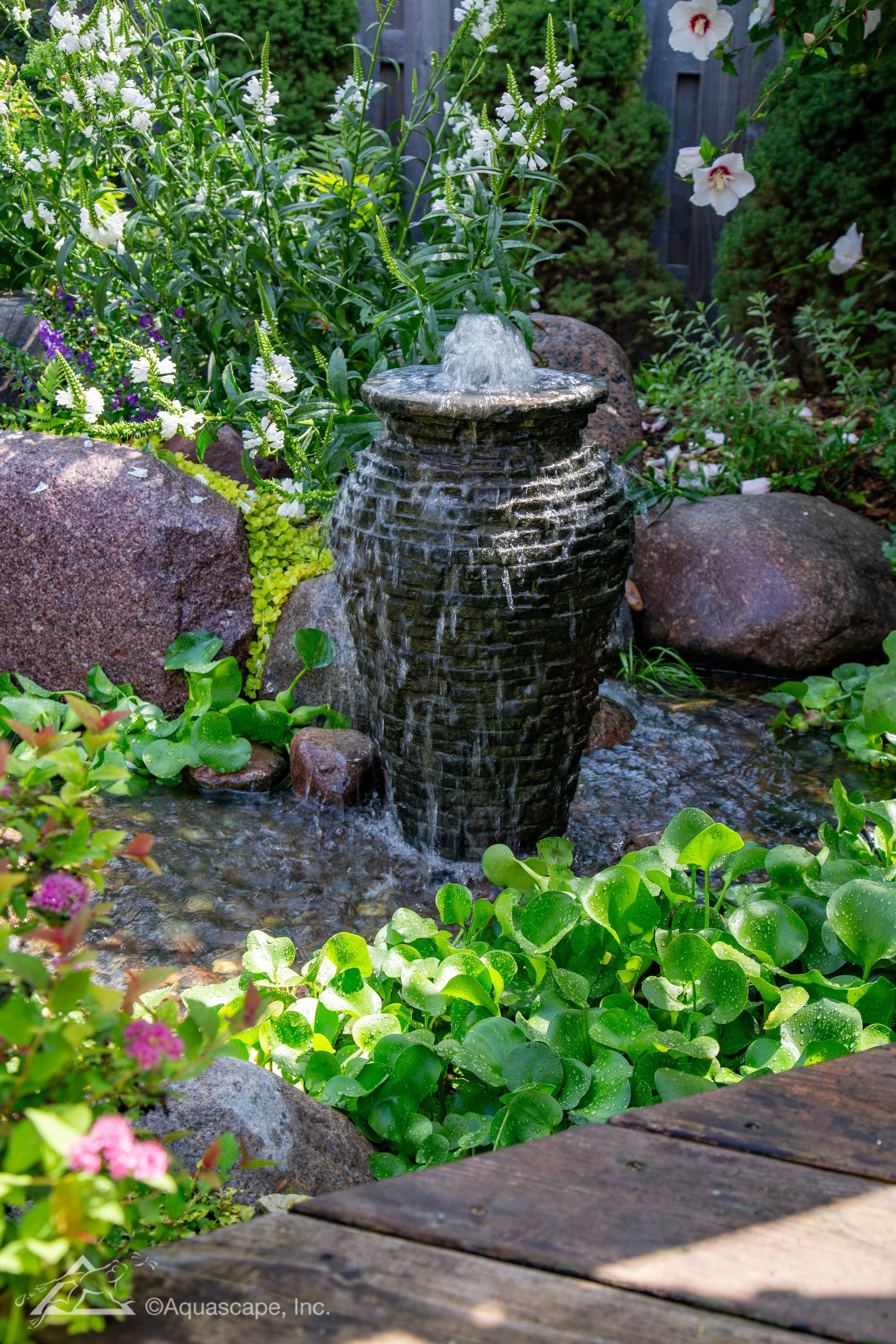 Stacked slate urn fountain with water cascading down its textured surface, surrounded by natural stone, aquatic plants, and lush garden foliage.