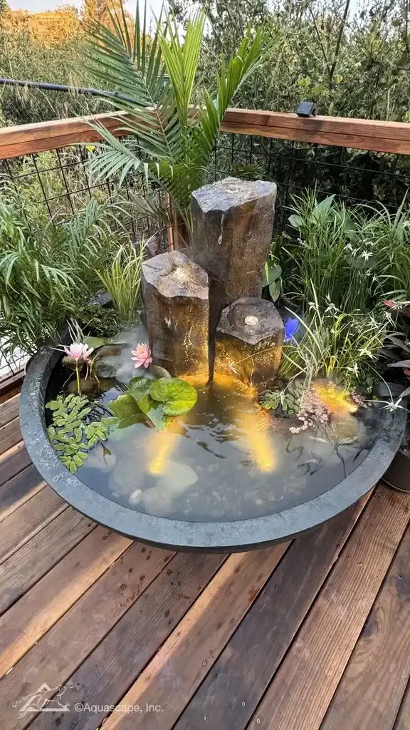 A small pond with water lilies and floating plants, featuring three stacked rocks with waterfall at the center, surrounded by tropical plants on a wooden deck.