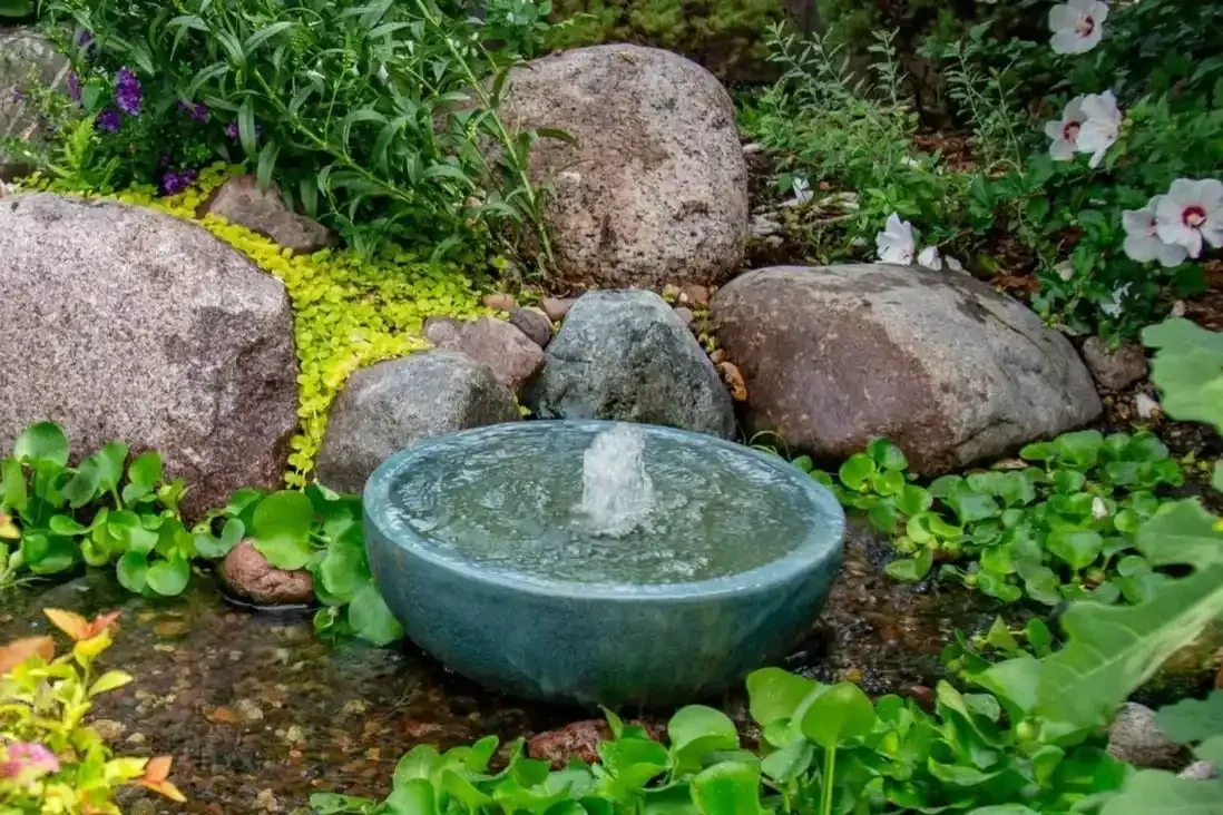 A small water fountain in a garden surrounded by green plants, colorful flowers, rocks, and pebbles.